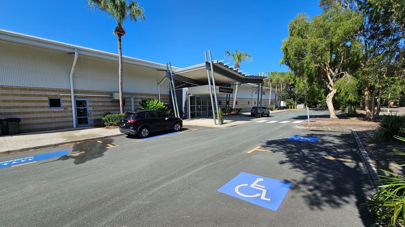 A modern building with a striped exterior and a large entrance canopy supported by angled metal beams. On the left, a black car is parked near a small palm tree. The foreground features a marked disabled parking space on the asphalt. On the right, there are several large trees and landscaped greenery. The sky is clear and blue.