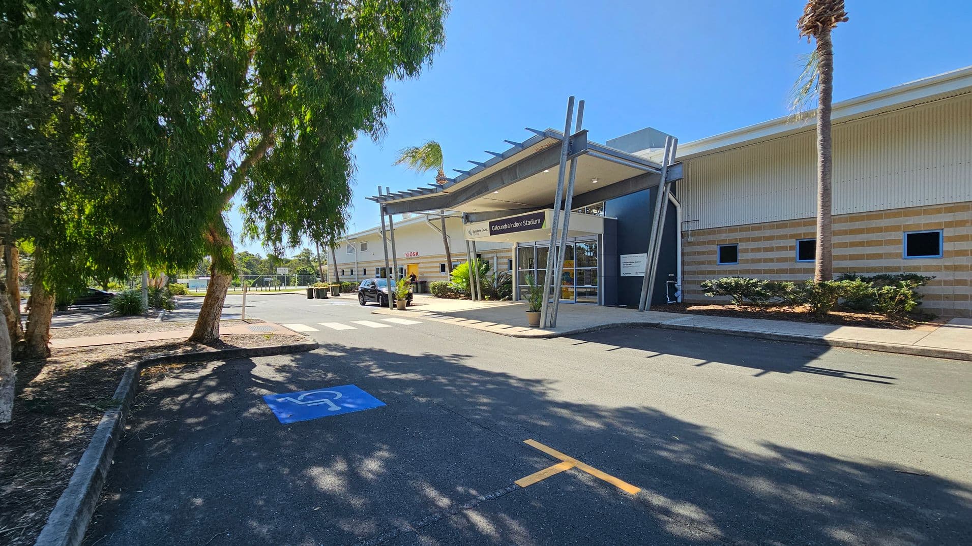 A large building with a modern entrance labelled "Caloundra Indoor Stadium" is on the right. The structure has a metal canopy supported by angled beams. A car is parked near the entrance. On the left, there are several large trees casting shadows on the ground. The pavement has a blue disabled parking symbol. The building's exterior is a mix of beige and cream with small windows. A palm tree is visible near the entrance.