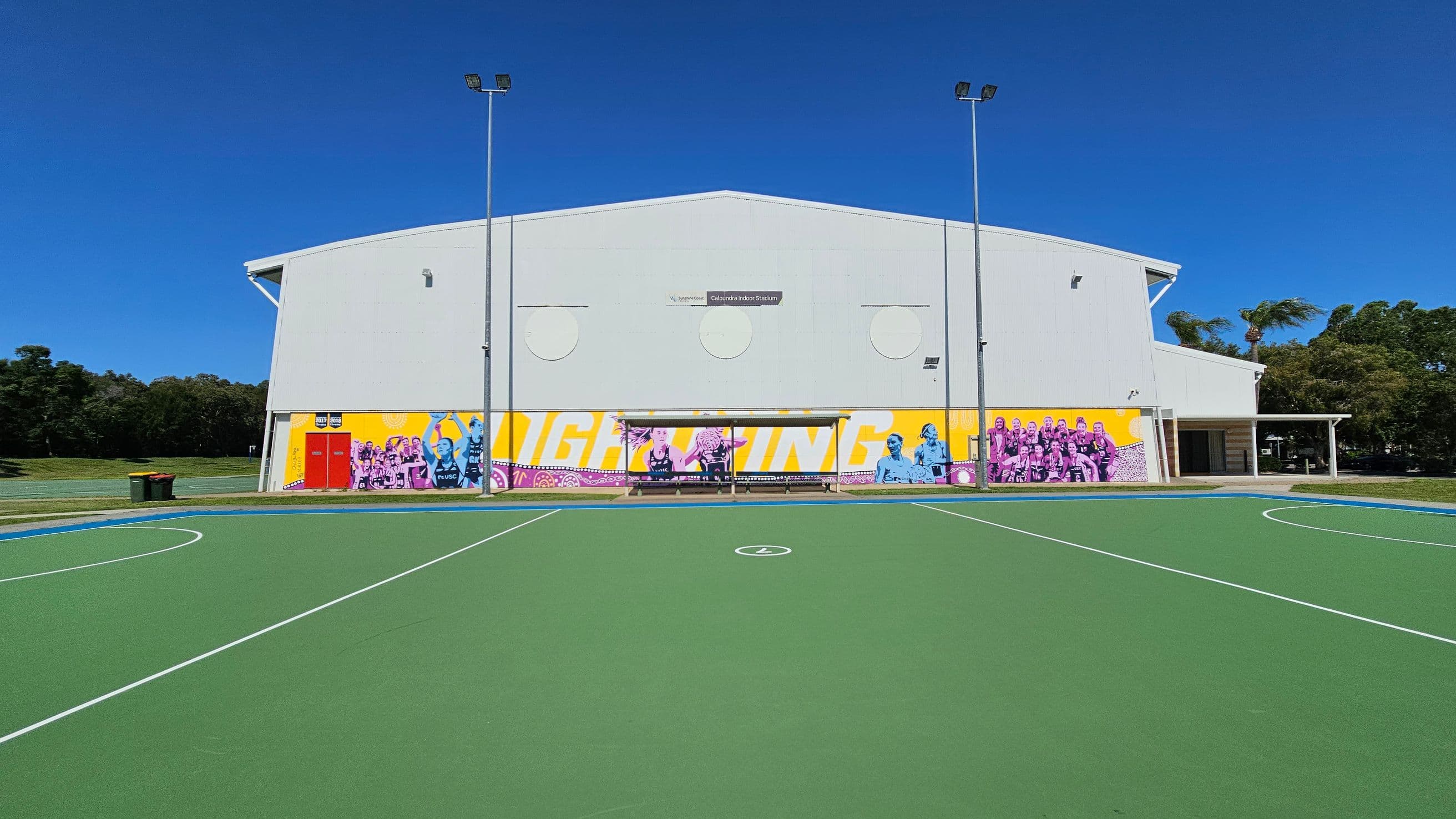 A sports court with a green surface and white lines in the foreground. In the background, a large white building with colourful murals featuring people and text. The building has a sign reading "Caloundra Indoor Stadium" and is flanked by tall light poles. To the left, there are green bins and grass, while trees are visible on both sides.