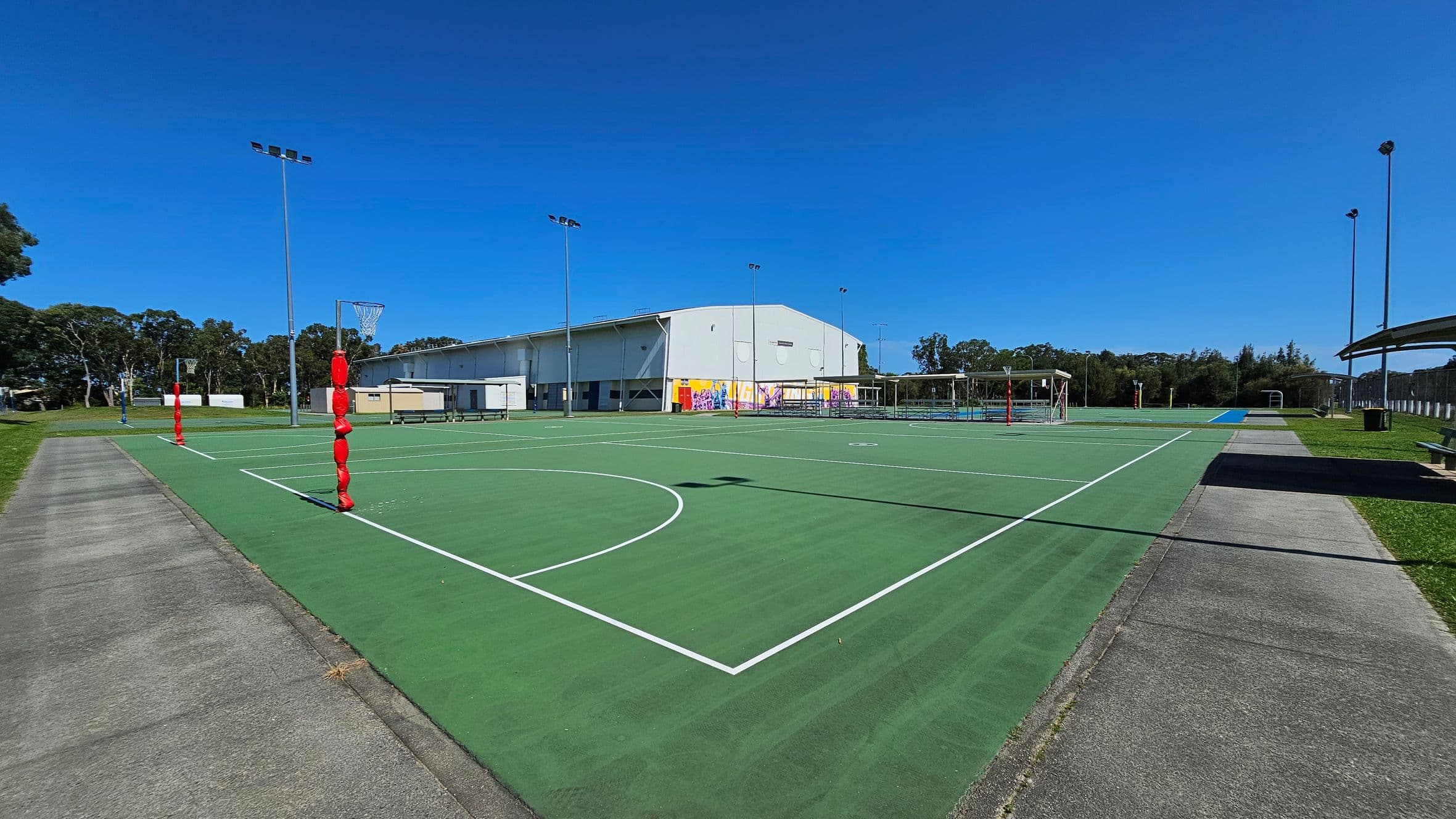 A green outdoor netball court with white lines is in the centre. Red padding covers the goal posts. On the left, a concrete path runs alongside the court, bordered by grass. In the background, a large building with colourful murals is visible. Several tall floodlights surround the area. On the right, there are more courts and a covered seating area. Trees line the horizon.