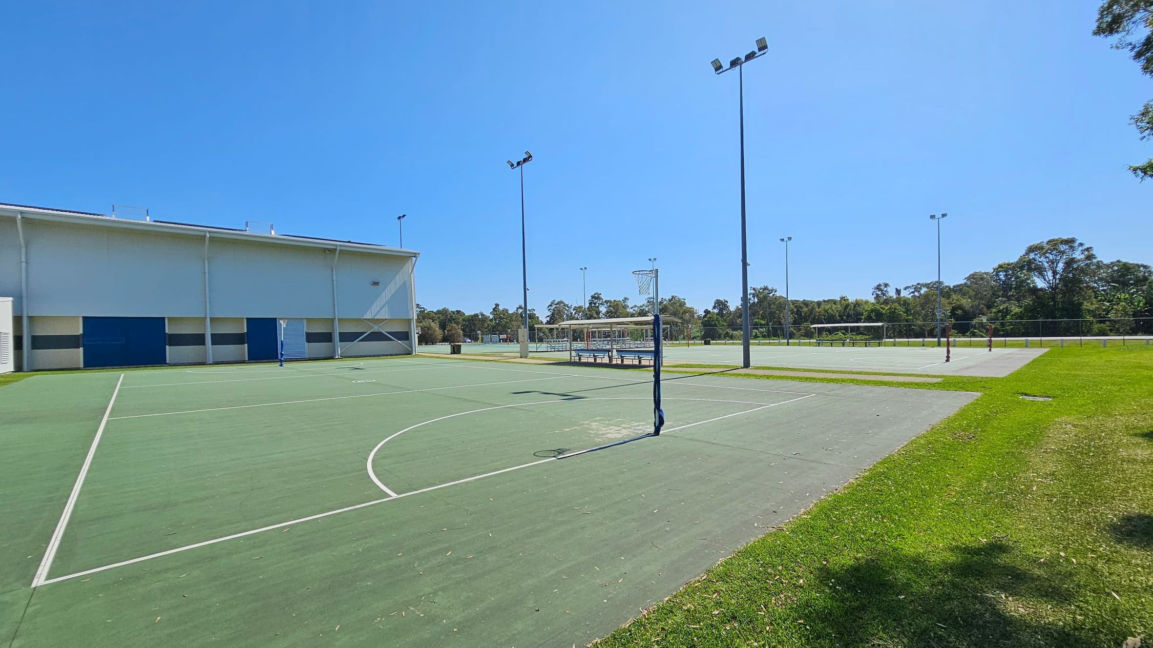 Outdoor sports courts with green surfaces and white lines. On the left, a large building with a white and blue exterior. In the centre, a netball hoop and tall floodlights. To the right, more courts and a grassy area. Trees and a fence line the background.