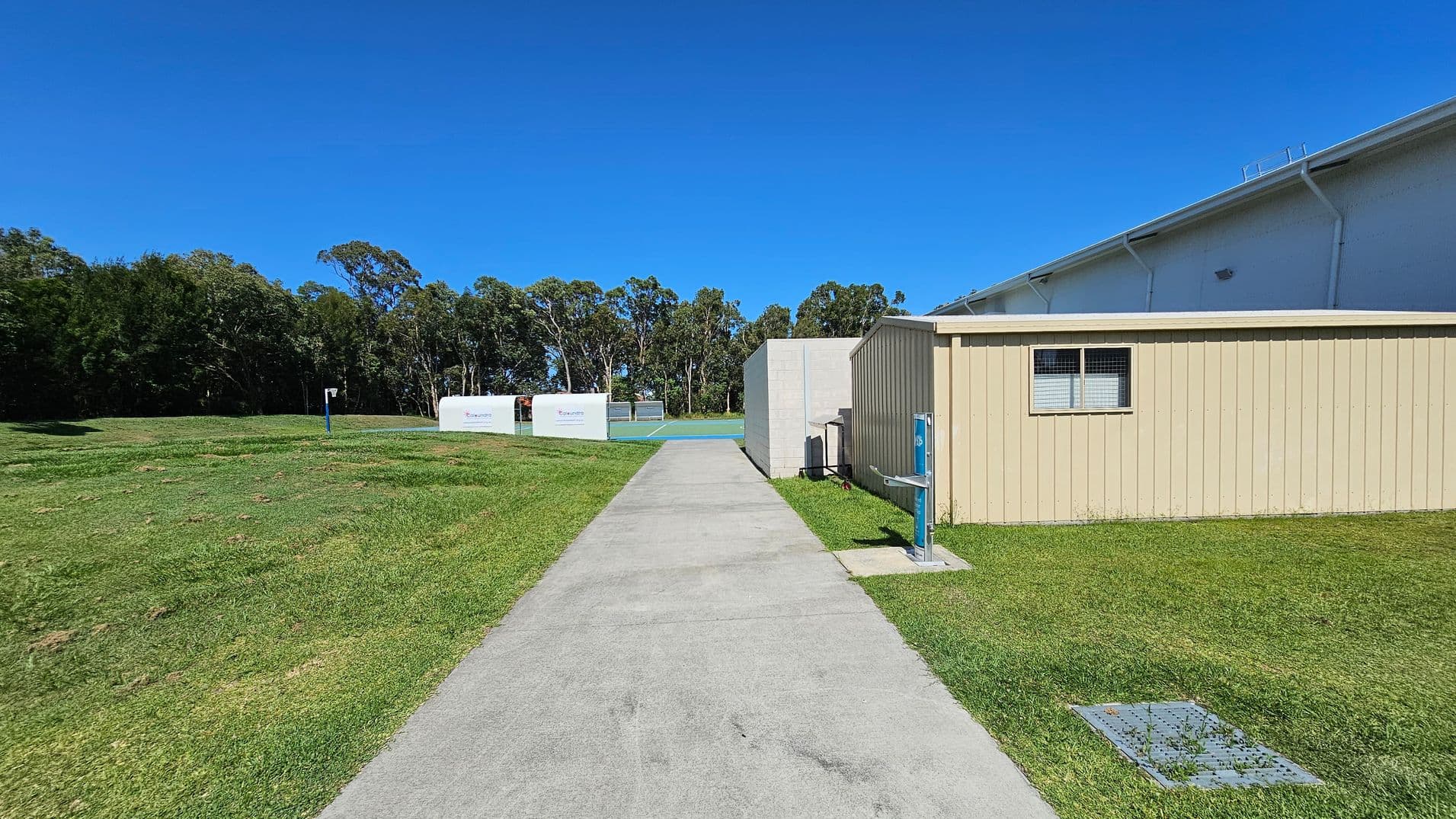 A concrete path runs through the centre, flanked by green grass. On the right, there's a beige building with a small window. To the left, open grassy fields extend towards a line of trees in the background. In the distance, there are two white structures with signage. The sky is clear and blue.