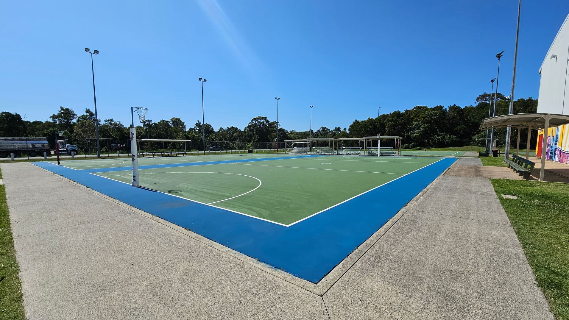 A netball court with a green playing surface and a blue border. On the left, a concrete path runs alongside the court, with a truck visible in the background near some trees. On the right, there is a covered seating area with benches and a building with colourful murals. Tall floodlights surround the court, and trees line the background.