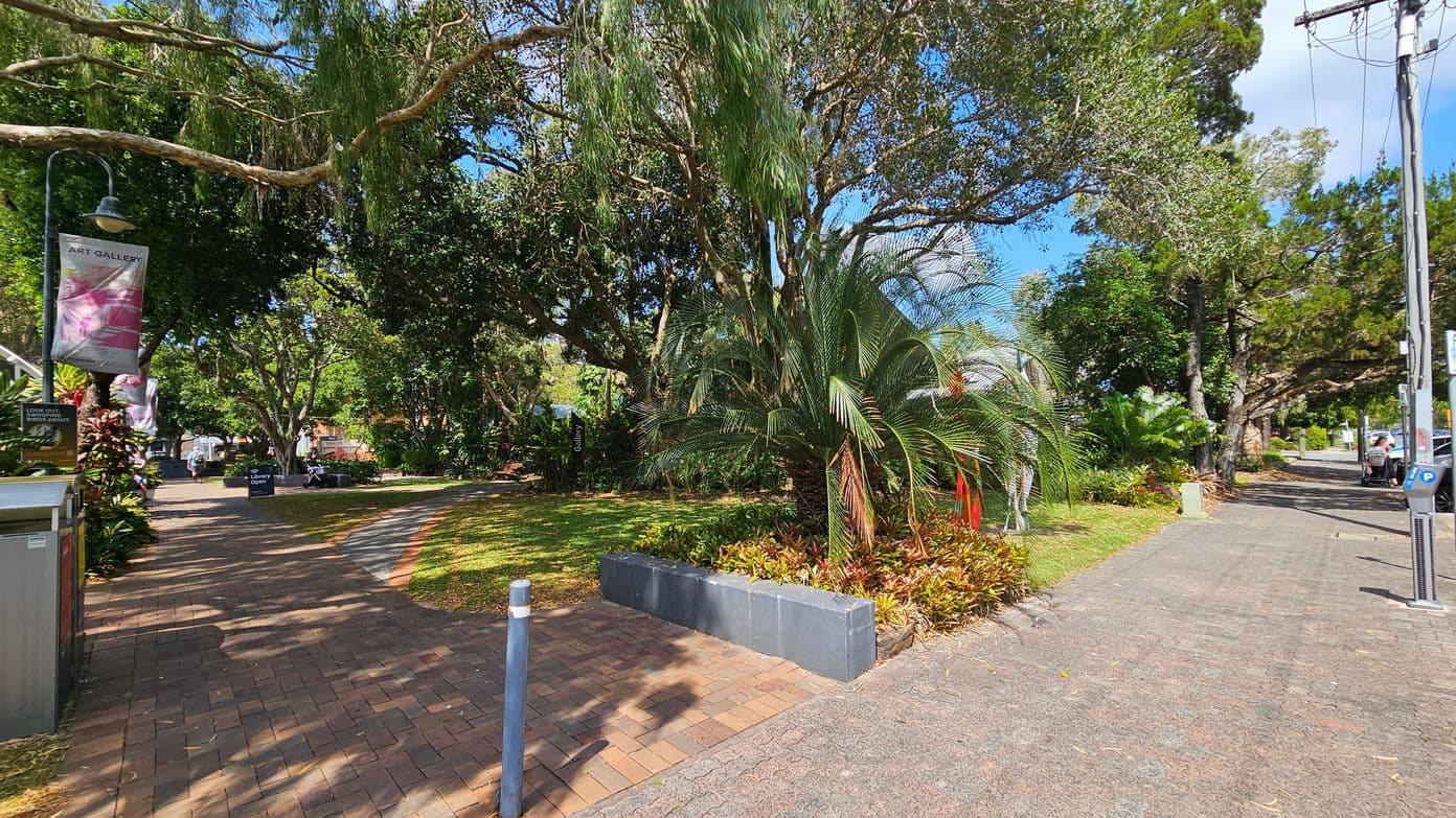 A brick pathway on the left leads through a park with lush greenery and large trees providing shade. A sign for an art gallery is visible, along with a lamp post. On the right, a concrete pavement runs parallel, bordered by more trees and plants. A parking meter and utility pole are present. In the background, people can be seen walking and sitting, enjoying the park's atmosphere.