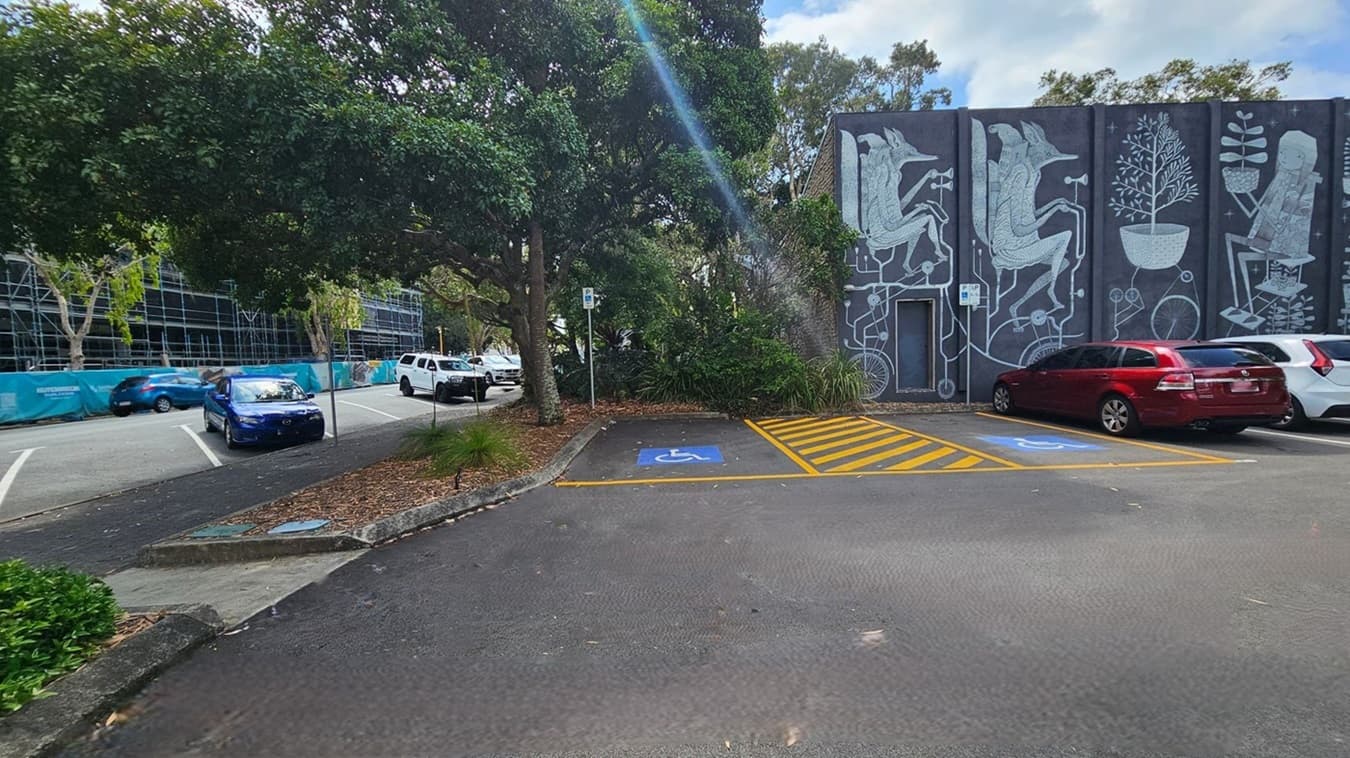 A car park with designated disabled parking spaces marked in blue and yellow on the right. A mural with abstract figures and plants is painted on the building wall in the background. On the left, a row of parked cars, including a blue car, is partially shaded by large trees. A construction site with scaffolding and a blue fence is visible further left. The ground is covered with scattered leaves.