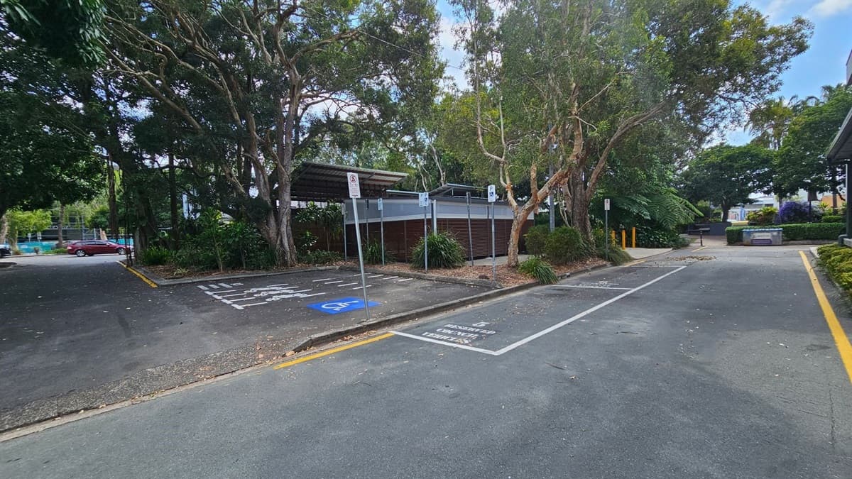 A parking area with a designated disabled parking space marked in blue on the left. Large trees with dense foliage surround the area, providing shade. A small building with a metal roof is in the centre, partially obscured by trees. A pathway leads to the right, bordered by greenery. A red car is parked in the distance on the left. The ground is covered with scattered leaves and debris. Signs are posted near the parking spaces.