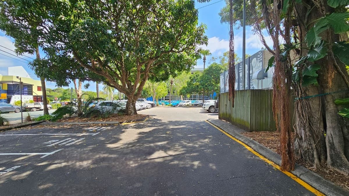 A car park with several vehicles is surrounded by trees. On the left, a large tree provides shade, with a yellow and white building visible in the background. On the right, a wooden fence with a mural runs alongside the pavement, with more trees and foliage. The ground is covered with fallen leaves, and the area is well-lit by sunlight filtering through the trees.