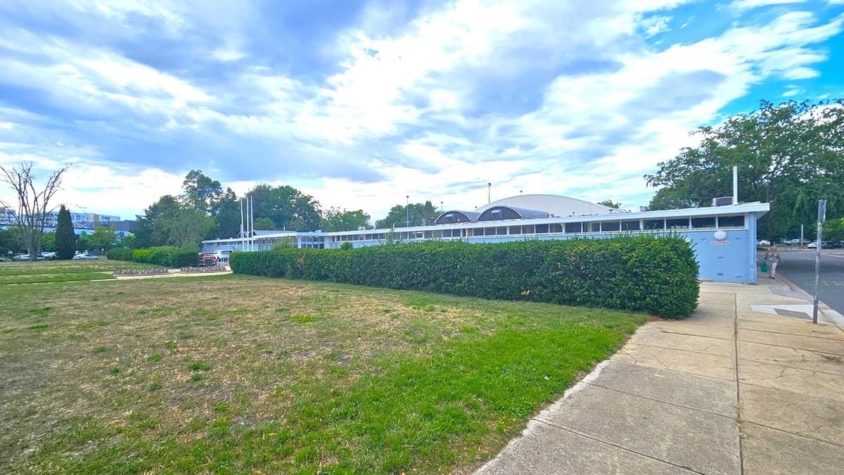 To the left, an expansive grassy parkland extends. On the right, a brick pathway and a road with a step-free entrance are present. Behind stands a blue concrete building with a dome-shaped roof. Trimmed ornamental shrubs border the building's front. Towards the far left, a concrete bike rack is positioned. The area is surrounded by trees.