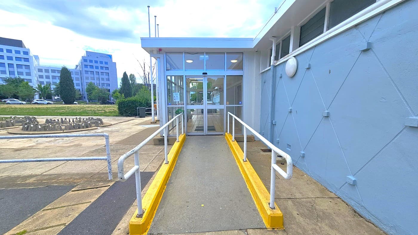 On the left, a concrete bike rack is positioned. A ramp with handrails providing access to the entrance is in the middle. To the right, a blue concrete wall with a diamond-shaped design is present. The building entrance features automatic glass doors. On the far left, there are trees, tall commercial buildings, and green grass visible.