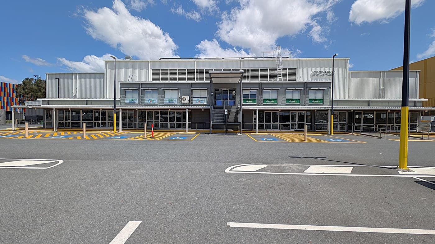 A large, modern building with a grey facade and multiple glass doors at the front. The upper section has several windows with green awnings. The foreground features a spacious car park with marked parking spaces, including accessible spots with blue and yellow markings. A colourful structure is partially visible on the left side, and there are several tall lamp posts around the area. The ground is paved with asphalt.