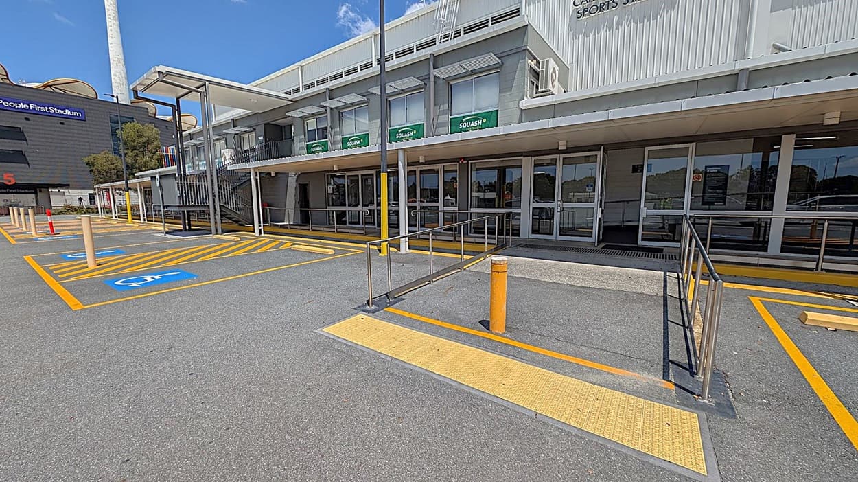 A parking area with designated disabled parking spaces marked in blue and yellow lines on the left. A ramp with metal railings leads to the entrance of a building on the right, which has large glass doors and windows with "SQUASH" signs. The building is grey with a flat roof and a covered walkway. A tall structure with a chimney is visible in the background on the left. The ground is paved with asphalt.