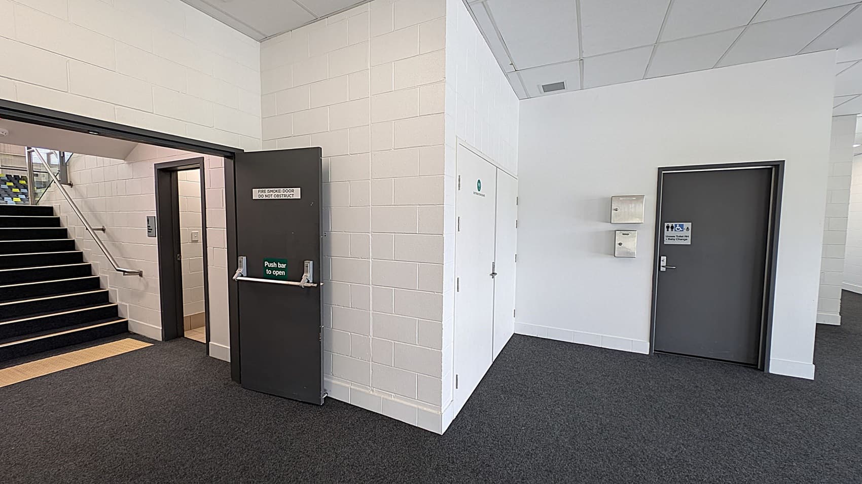 A corridor with a dark grey fire door on the left, labelled "Fire Smoke Door Do Not Obstruct," leading to a staircase with a metal handrail. The floor is carpeted in dark grey. On the right, there is a grey door with a sign indicating accessible toilets. Above this door are two wall-mounted boxes. The walls are painted white, and the ceiling is made of white tiles.