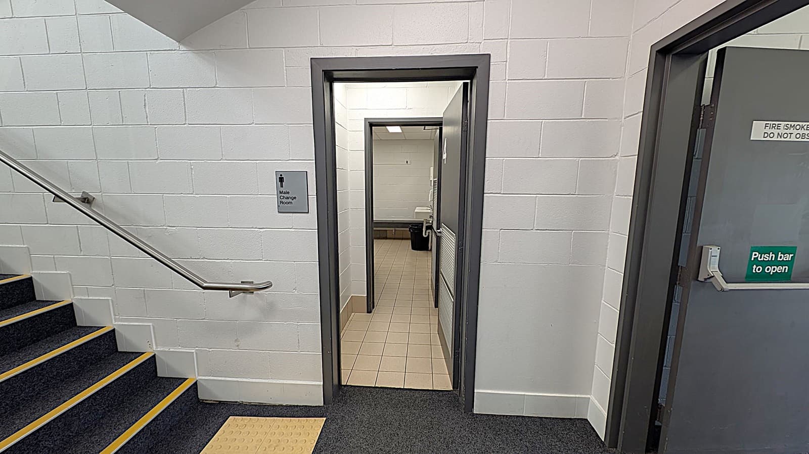 A corridor with a white brick wall on the left, featuring a staircase with a metal handrail and black steps with yellow edges. In the centre, there's an open door leading to a male changing room, with a sign on the wall. The floor inside is tiled. On the right, a grey door with a "Push bar to open" sign. The ceiling has fluorescent lights.