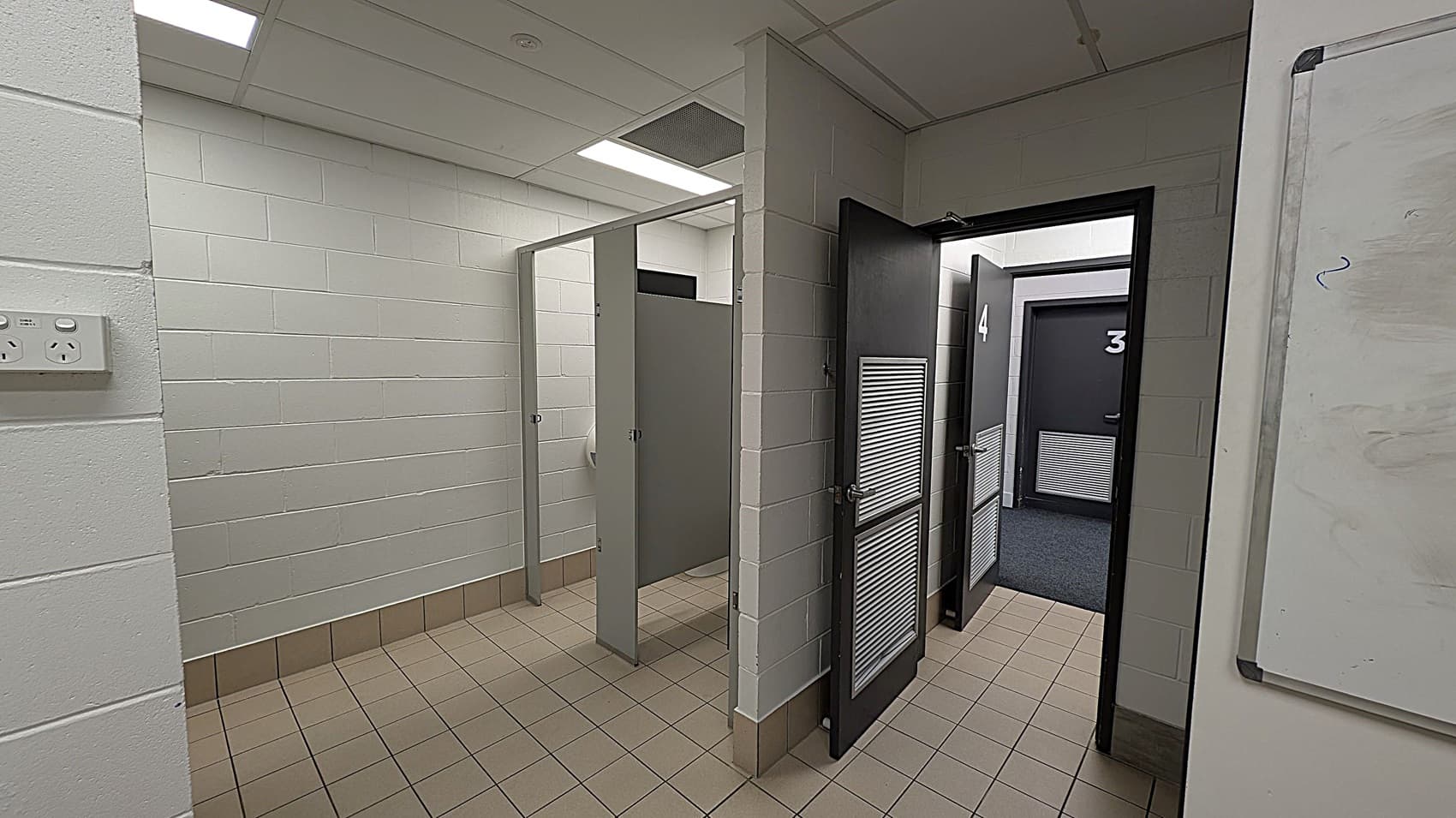 A corridor with tiled flooring and white brick walls. On the left, there are two open cubicles with grey doors. On the right, two closed doors numbered 3 and 4, each with a vent panel. The ceiling is fitted with fluorescent lights. A whiteboard is partially visible on the far right wall.