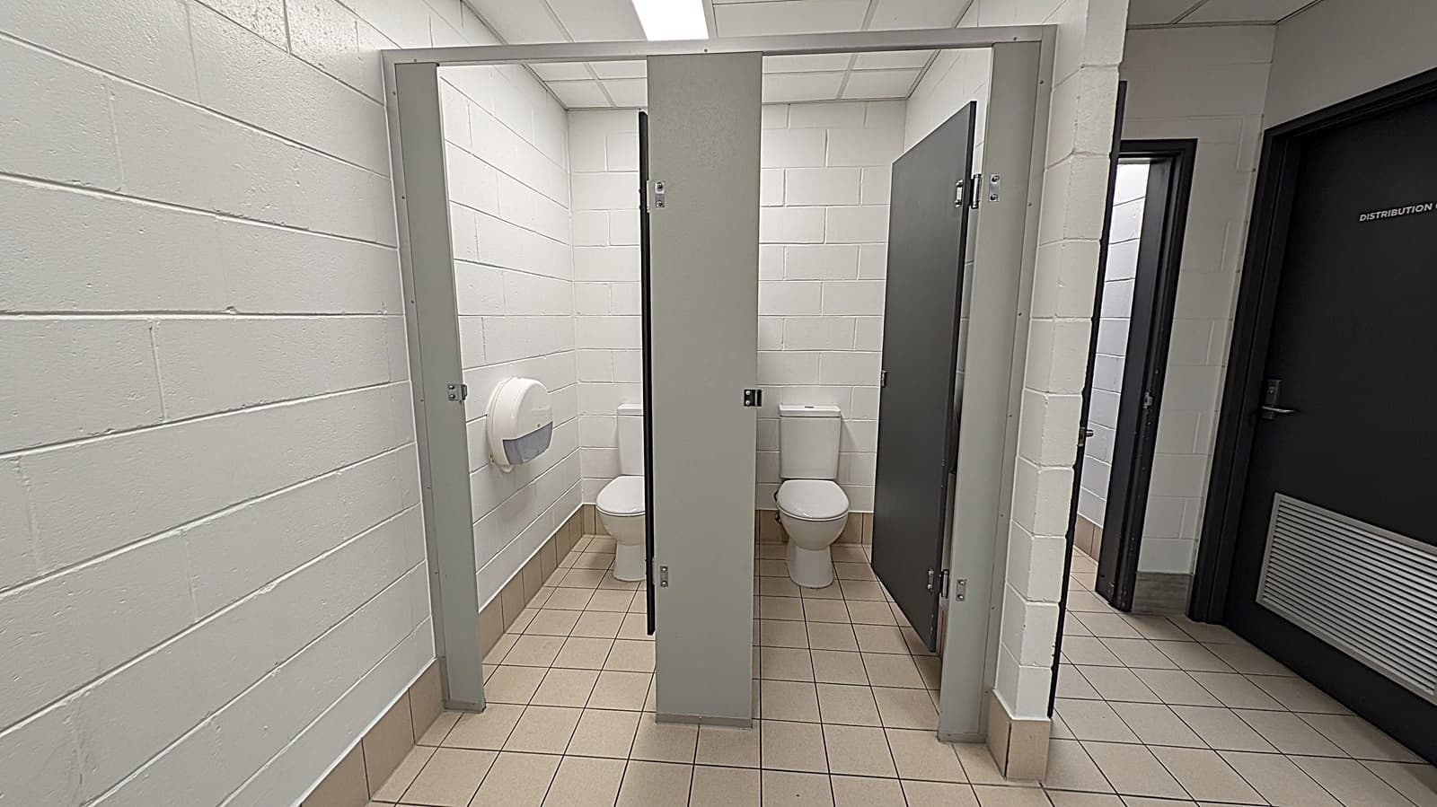 A public restroom with two toilet stalls in the centre, each with a white toilet and grey doors. The walls are made of white tiles, and there is a toilet paper dispenser on the left wall. The floor is covered with beige tiles. On the right, there is a dark door labelled "DISTRIBUTION ROOM". The ceiling has fluorescent lighting.
