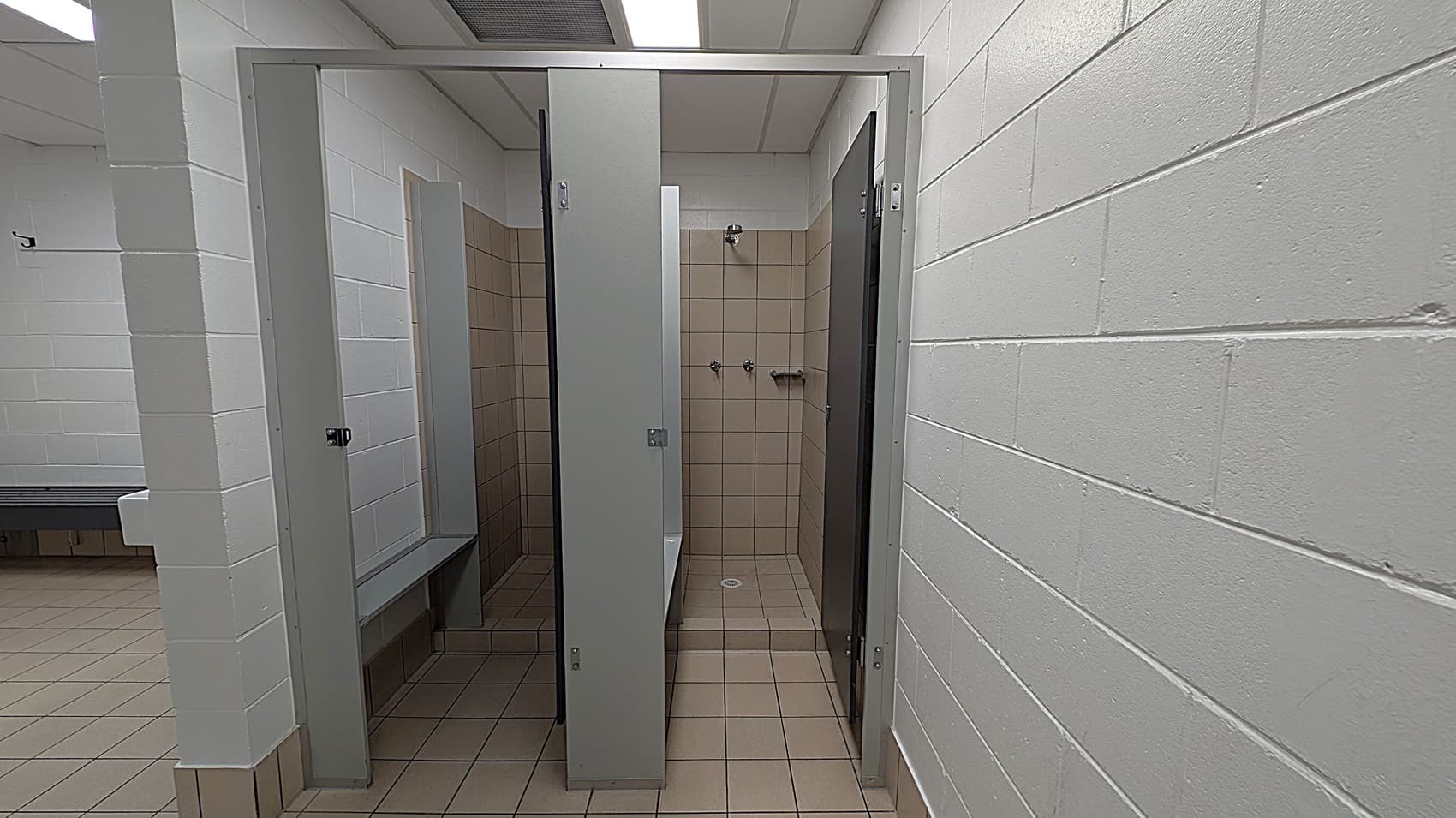 A public shower area with beige tiled walls and floor. Two showerheads and a small shelf are visible in the middle. The shower is enclosed by grey partitions with a door slightly ajar. The surrounding walls are white-painted brick. To the left, a glimpse of a green-tiled floor and a bench can be seen. The ceiling is white with fluorescent lighting.