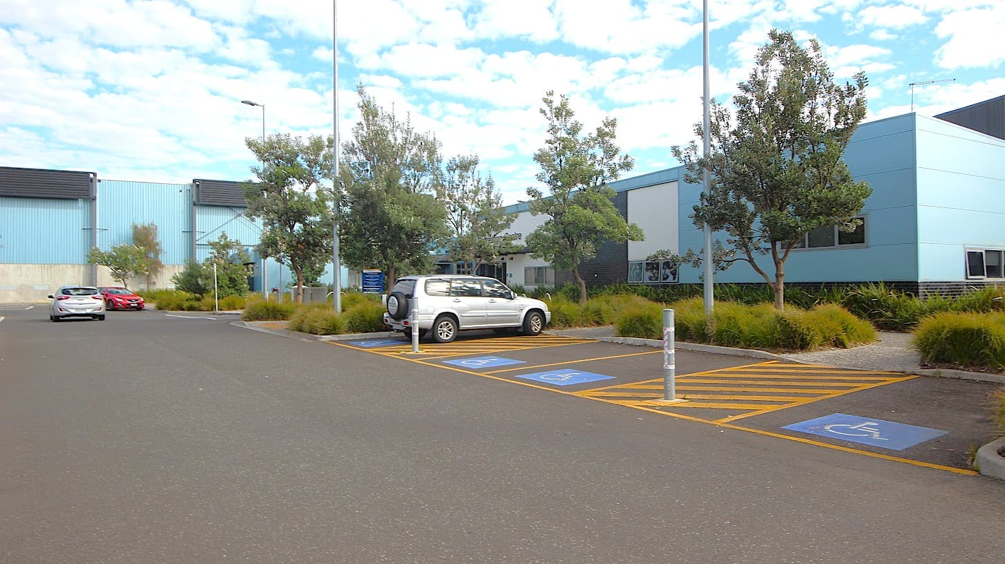 In the foreground, there is a large, paved parking area with multiple spaces marked, including two designated for disabled parking. A white vehicle is parked in one of these spaces. To the left, a grey car is visible further down the parking area. On the right, the building features blue cladding with several windows, surrounded by landscaped areas with small trees and shrubs. Several light poles are scattered throughout the parking lot, providing illumination.