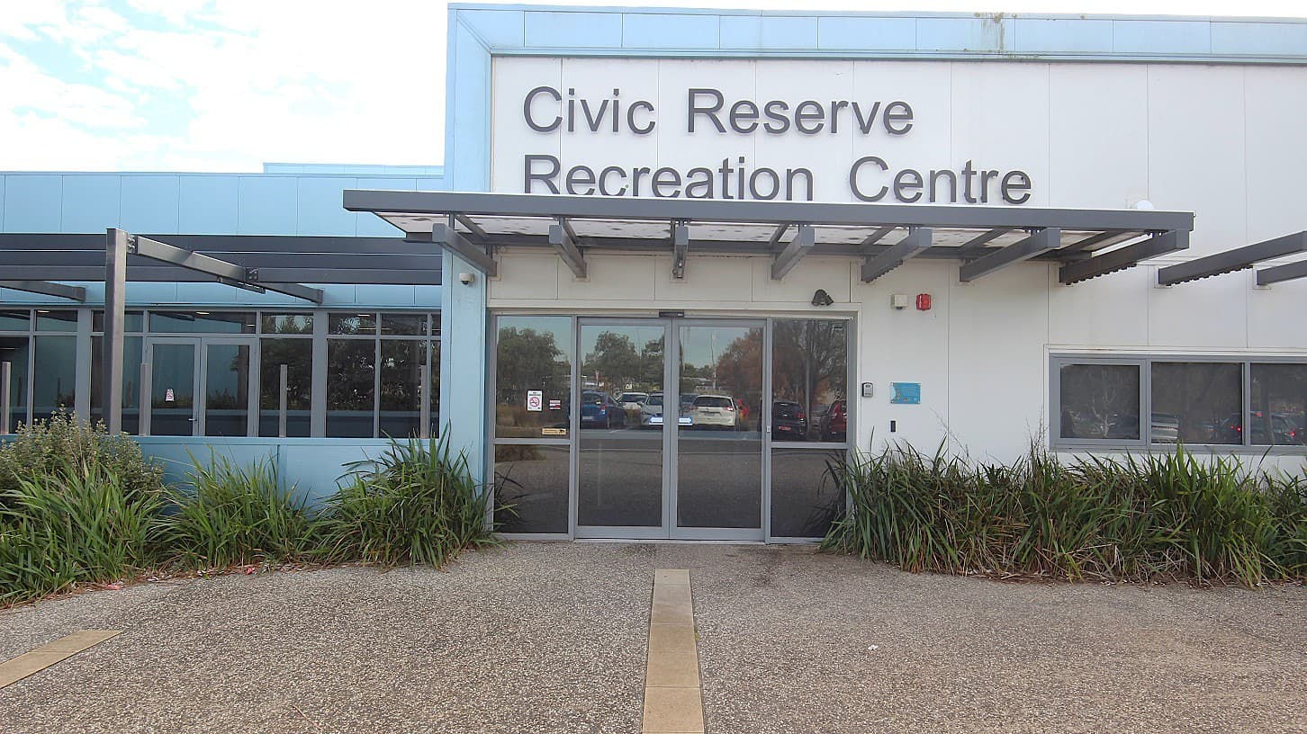 A closer look at the entrance glass doors with light grey painted edges and an overhang made of grey metal with a white membrane and transparent roofing at Civic Reserve Recreation Centre. Near the venue, there is a carpark where the glass reflects the cars parked in the middle. On the left door, there is a no-smoking sign. The name of the venue can be seen at the top. Covering the lower portion of the walls of the venue are plants with parallel venation.