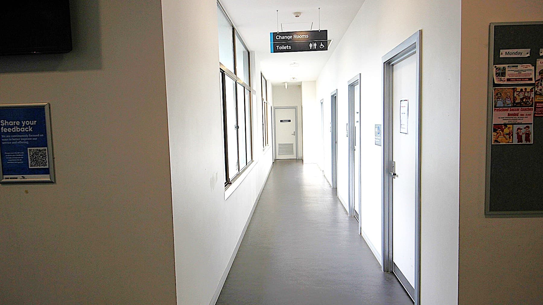 Hallway to the changing rooms and bathrooms at Civic Reserve Recreation Centre. The walls are white, and the floor is grey. On the opposite side of the 4 doors on the right with sky-blue painted edges are glass windows. Hanging on the ceiling is a small black signboard for Change rooms and Toilets.