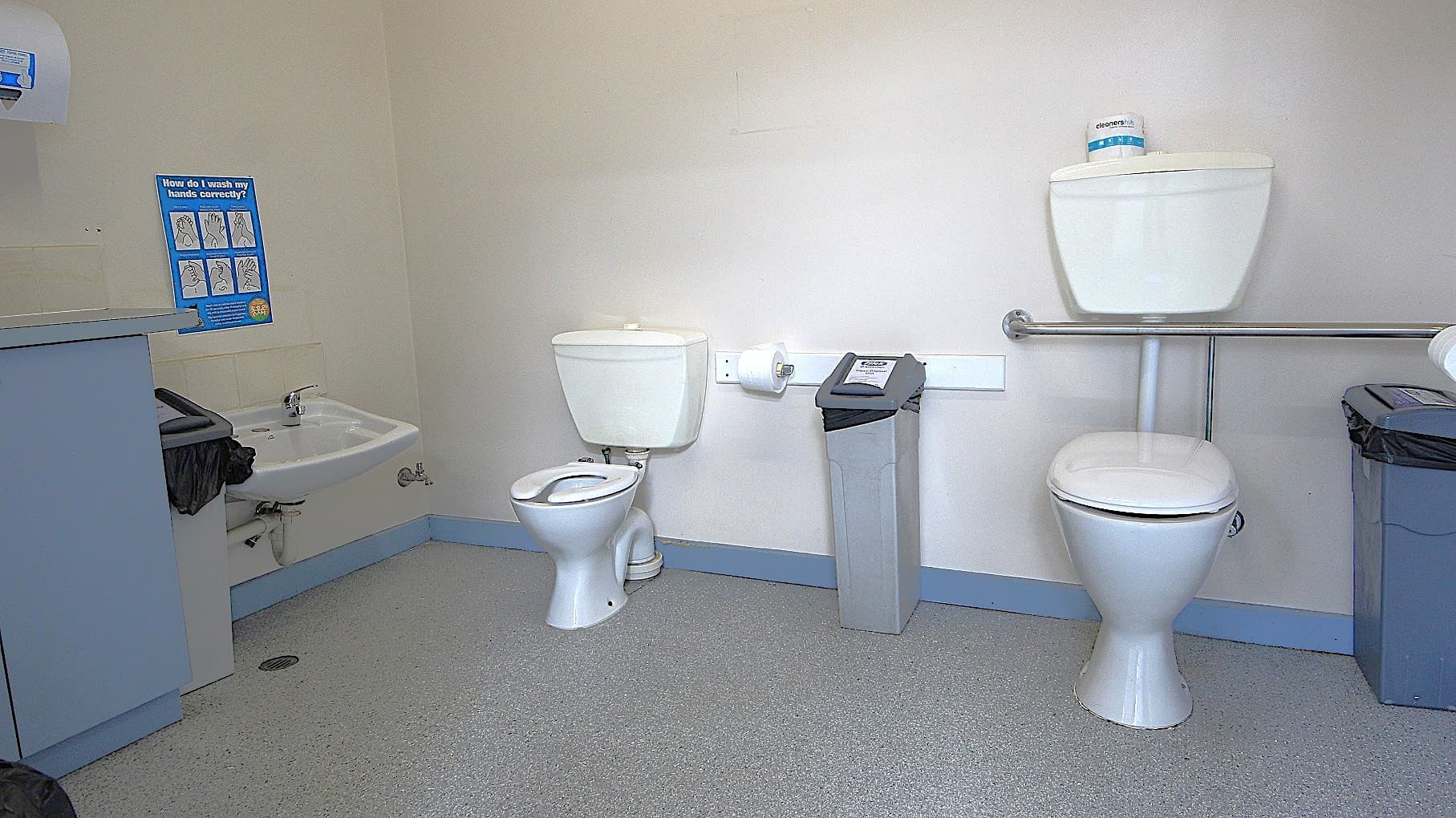 Inside the accessible bathroom at Civic Reserve Recreation Centre. On the left wall is a paper towel dispenser, a grey rubbish bin, a low-set wash basin, and a blue infographic about how to wash hands properly. In the middle are two toilet seats of different heights and sizes (left small; right big). Between them is a sanitary napkin bin and a toilet roll holder with toilet paper. The floor is granite, and the walls are painted white.