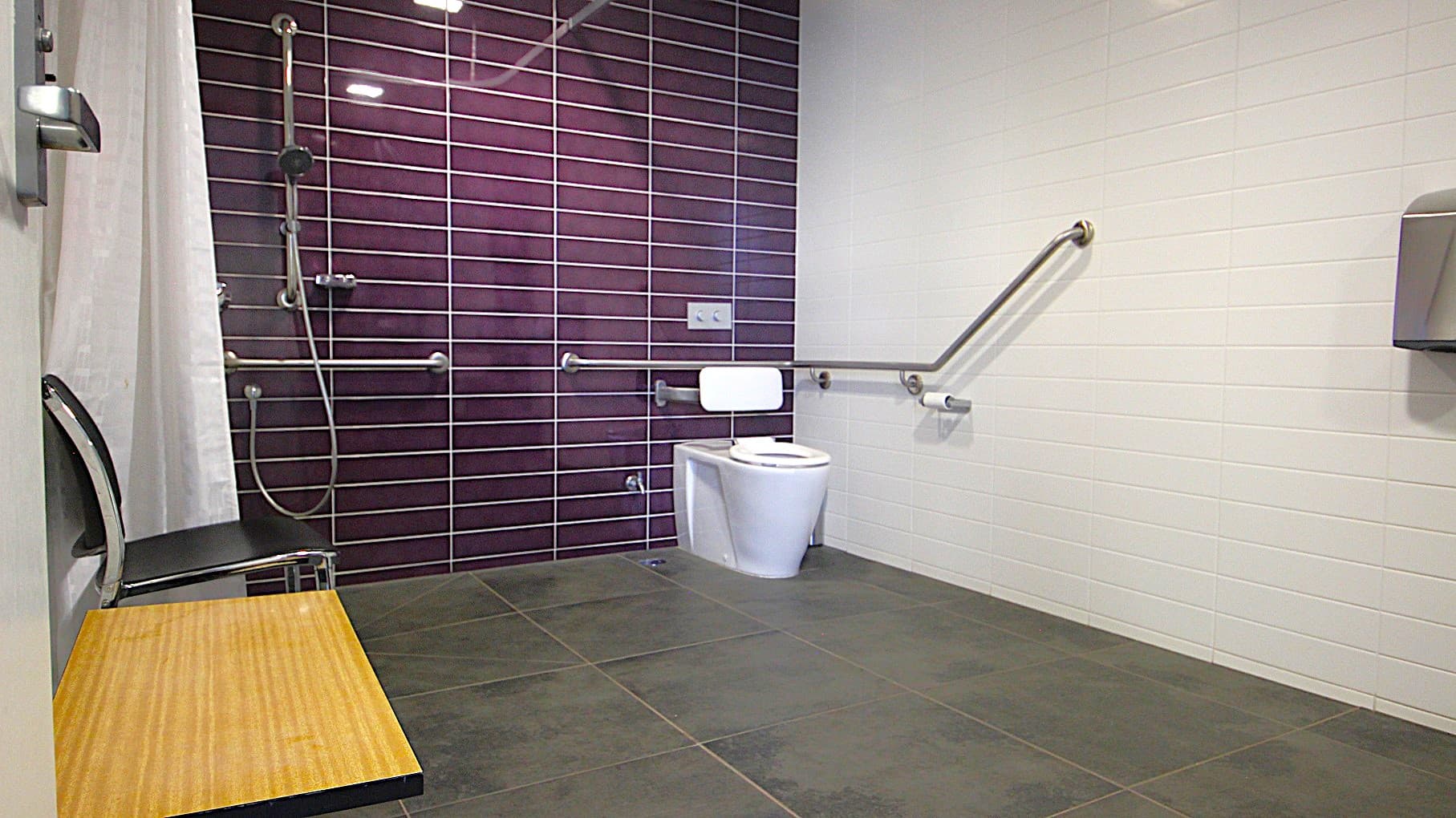 Image inside one of the accessible bathrooms at Civic Reserve Recreation Centre. On the left is a knee-high wooden table and a black chair next to it. There is a white shower curtain, a handheld showerhead, and grab bars situated on the purple-tiled wall. and to the right is a toilet. The bathroom has a grey-tiled floor and a white-painted brick wall.