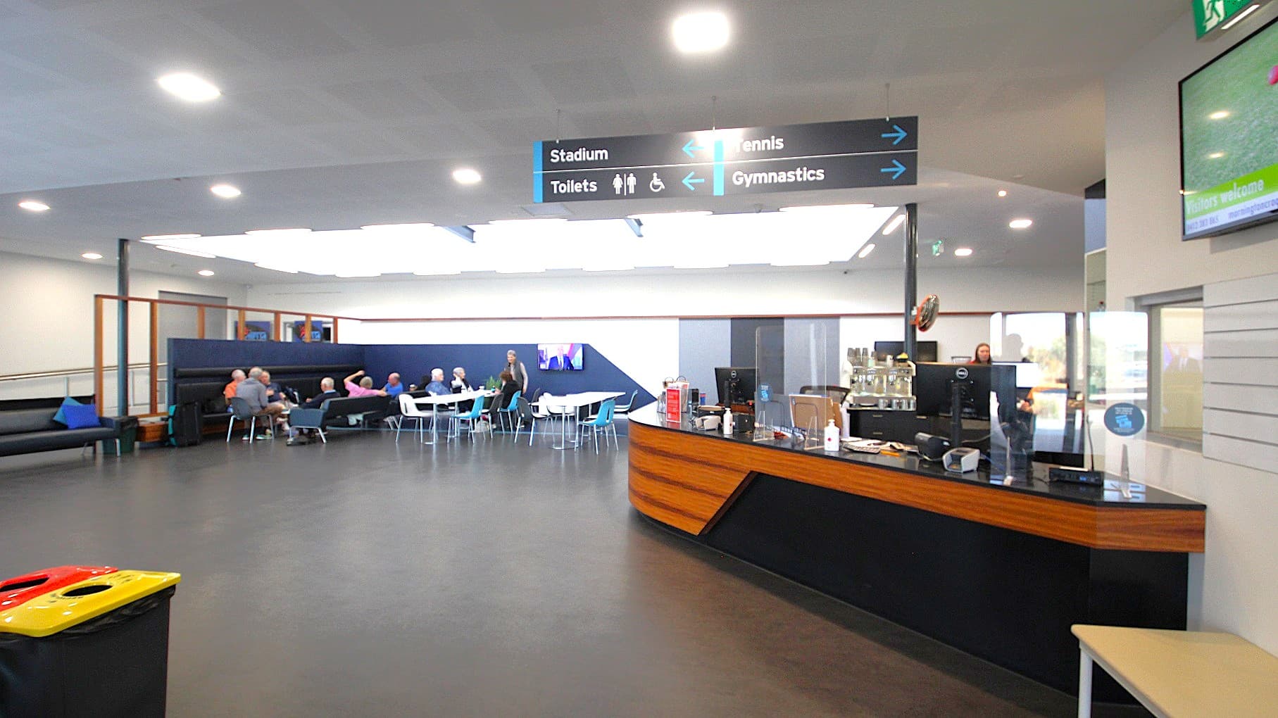 The first section inside the Civic Reserve Recreation Centre is a black and walnut counter of the café and seating area occupied by adults enjoying themselves while chatting. Lighting the seating area is a skylight allowing natural light to filter in addition to fluorescent strips that surround this rectangular skylight. There is black signage indicating the location of the Stadium, Toilets, Tennis, and Gymnasium.