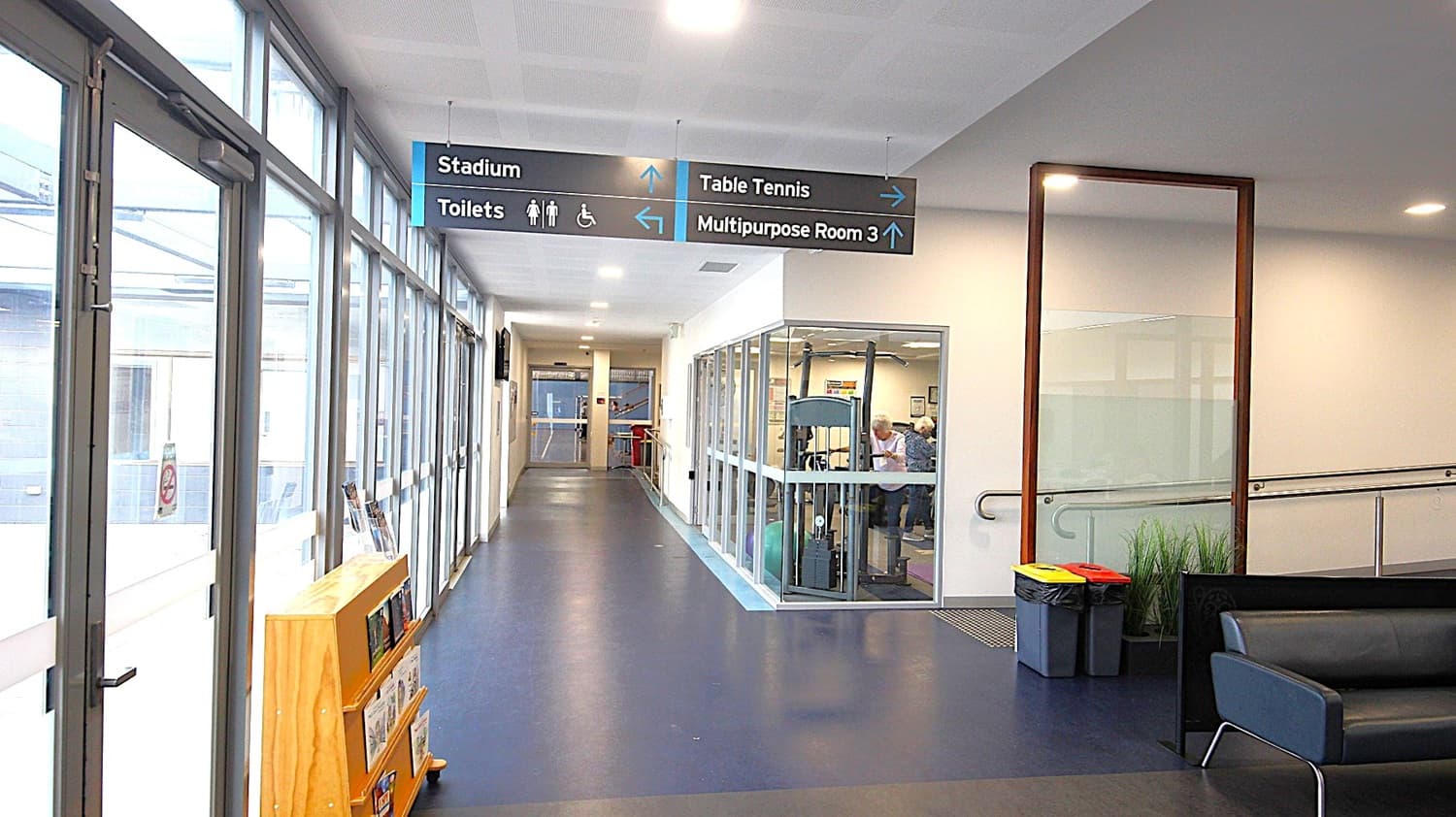 Hallway on the left side of the main entrance doors. The image shows a wooden brown cabinet behind the glass doors on the left, a low black sofa chair, and black rubbish bins with red and yellow lids at the start of the entrance ramp on the right. Looking straight ahead, this hallway has a grey floor, a room to the right filled with adults using exercising equipment, and hand rails on the white wall.