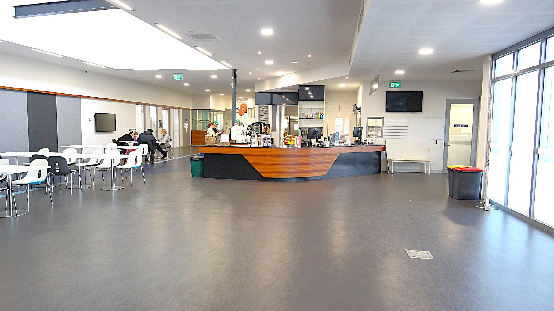 The image shows the side of the black and walnut café counter and its surrounding area. On the right side is a green fire exit sign. Below it is a television mounted on the upper corner of the wall, and next is the “Staff only” white, single, glass door. On the left is a seating area with white chairs and tables that have steel legs and frame.