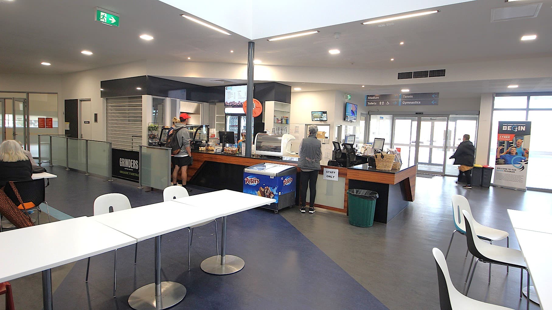 At the café counter is a black cash register, a glass display cabinet with mouth-watering pastries, and small, well-designed flyers with aesthetic colors. There are cold drinks stored in a large fridge, and the menu can be seen on the digital screen mounted on the wall above it. Further to the left is a separate walkway by grey stanchions towards a white door room next to the café.