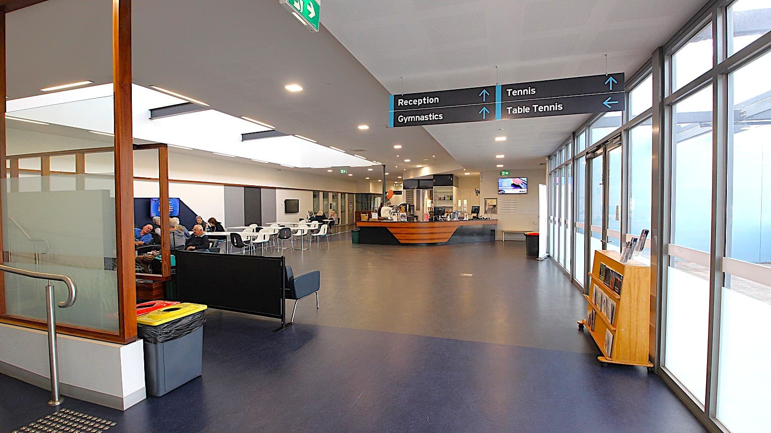 View of the café and seating area from the entrance ramp. The attractive feature of this floor is the skylight above the seating area of the café. The floor is painted in grey and black. The ceiling is white with small, square LED lights. Rubbish bins are placed randomly on this floor for better waste management, and directional signage is large enough to be seen.