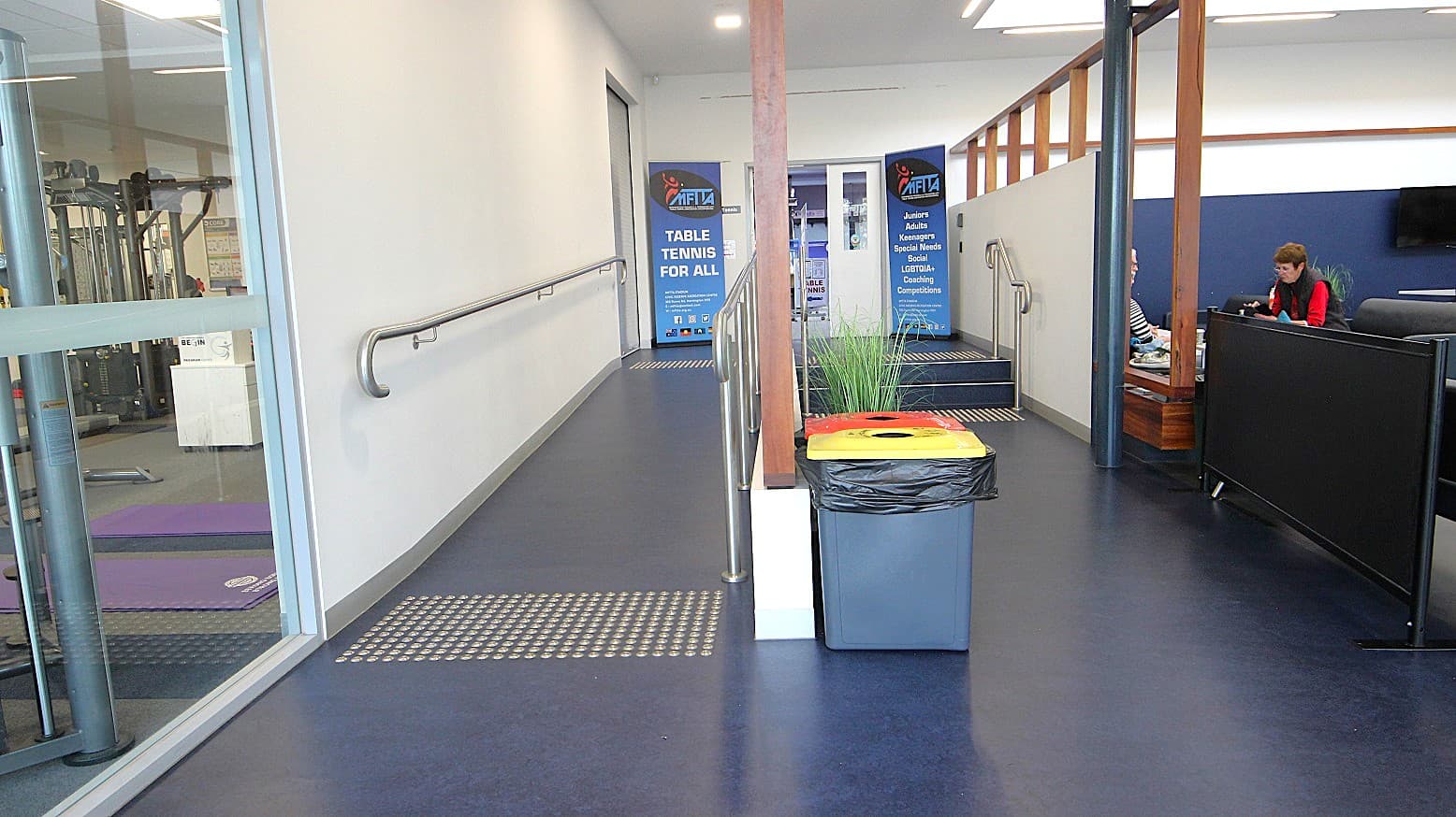 Entry to the table tennis area is via the entrance ramp and a two-step tier stairwell, both of which have handrails to assist individuals with mobility disabilities. The starting points of the ramp and stairwell are designed with tactile ground surface indicators. Potted plants and black rubbish bins with yellow and orange lids are located in front of the stairwell.