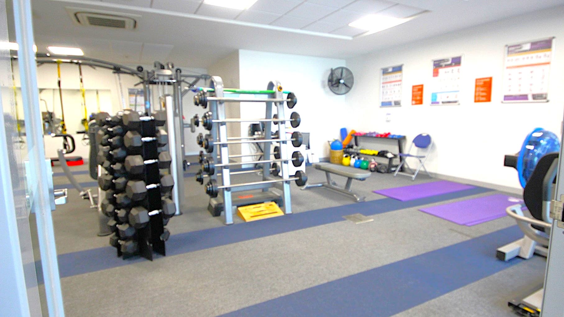 A gym with various equipment. On the left, there is a rack of dumbbells. In the centre, a weightlifting station with multiple barbells. On the right, exercise mats are laid out on the floor, with a rowing machine partially visible. The back wall has posters and a fan, with a bench holding kettlebells and medicine balls. The floor is carpeted in grey and blue stripes, and the ceiling has recessed lighting and an air conditioning unit.