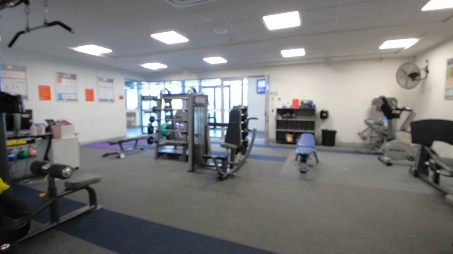 A gym with various exercise equipment. In the centre, there is a weight machine and a bench. To the right, a treadmill and a stationary bike are visible. On the left, there are more benches and weights. The walls have posters, and a fan is mounted on the right wall. The floor is carpeted with dark grey tiles, and the ceiling has recessed lighting. In the background, there are large windows and a glass door.