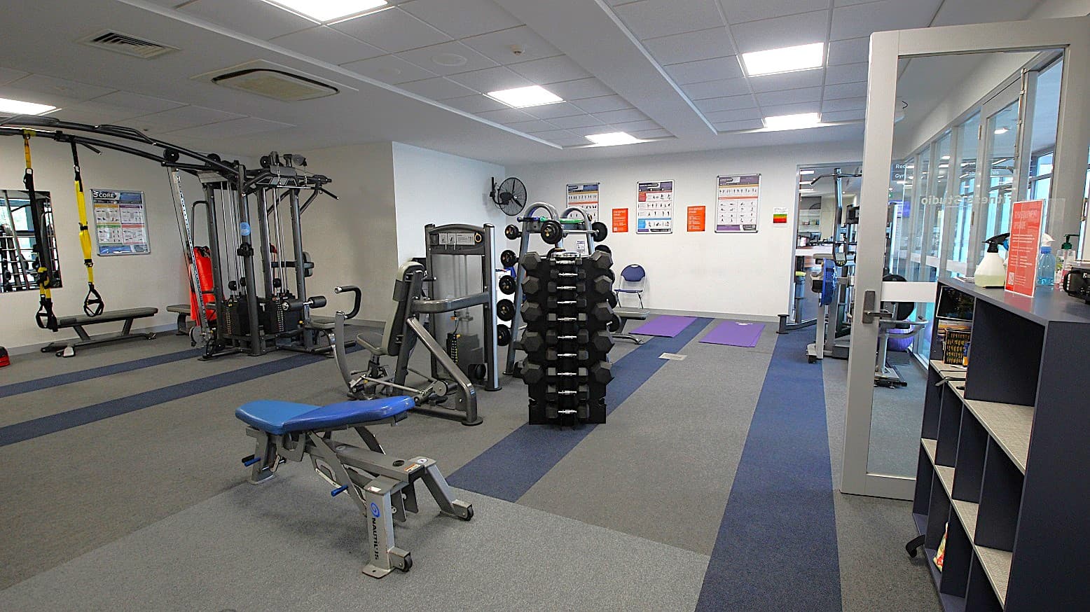 A gym with various exercise equipment. On the left, there is a multi-station machine and a bench with TRX straps hanging. In the centre, a rack of dumbbells is visible. The right side has a glass door and a shelving unit with books and cleaning supplies. The walls have instructional posters, and the floor is covered with grey and blue carpet tiles. The ceiling is fitted with fluorescent lights.