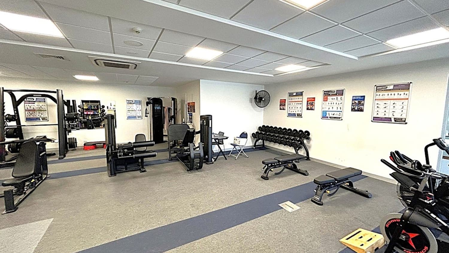 A gym with various exercise equipment. On the left, there are weight machines and a bench press. In the centre, a leg press machine and a seated bench are visible. On the right, a row of dumbbells is placed on a rack beneath wall-mounted exercise charts. A stationary bike is partially visible. The floor is carpeted in grey with a darker strip running through it. The ceiling is fitted with fluorescent lights and air conditioning vents. A wall fan is mounted near the dumbbells.