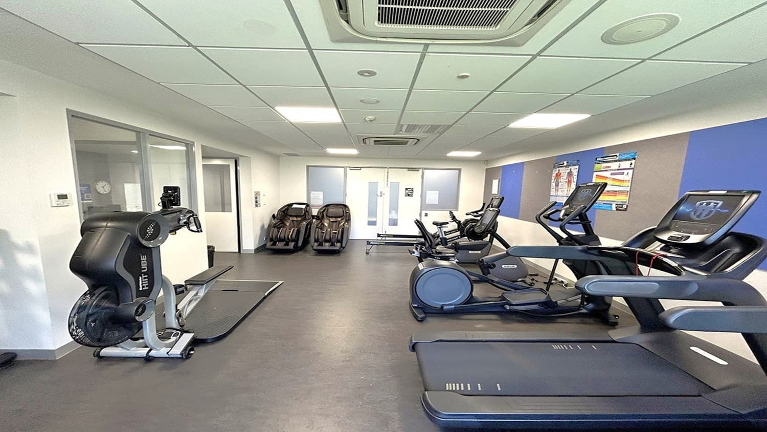 A gym room with exercise equipment. On the left, there's a FitLuge machine near a glass partition. In the middle, several cardio machines, including treadmills and exercise bikes, are lined up against the wall. On the right, two treadmills are visible. The walls have blue and beige panels with fitness posters. The floor is dark grey, and the ceiling is white with recessed lighting and air conditioning vents.