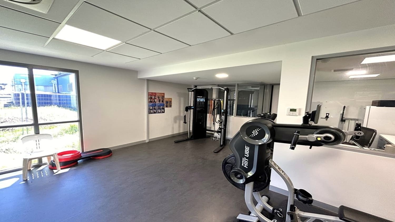 A gym room with exercise equipment. On the right, there's a large machine with "HIT UBE" written on it. In the centre, a multi-functional gym station is visible. On the left, a window allows natural light in, with a white chair and stacked red and black exercise steps nearby. The walls are light-coloured, and there are posters on the wall. The ceiling has recessed lighting, and the floor is covered with grey tiles.