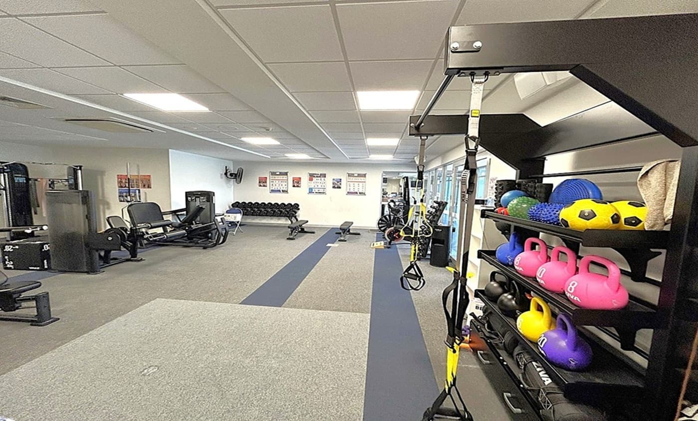 A gym interior with a variety of equipment. On the right, a rack holds colourful kettlebells, medicine balls, and other fitness gear. In the middle, a long strip of flooring leads to more equipment, including benches and weights. On the left, there are exercise machines and a row of dumbbells against the wall. The ceiling is fitted with fluorescent lights, and the floor is carpeted with different sections for various activities. Posters are visible on the walls.