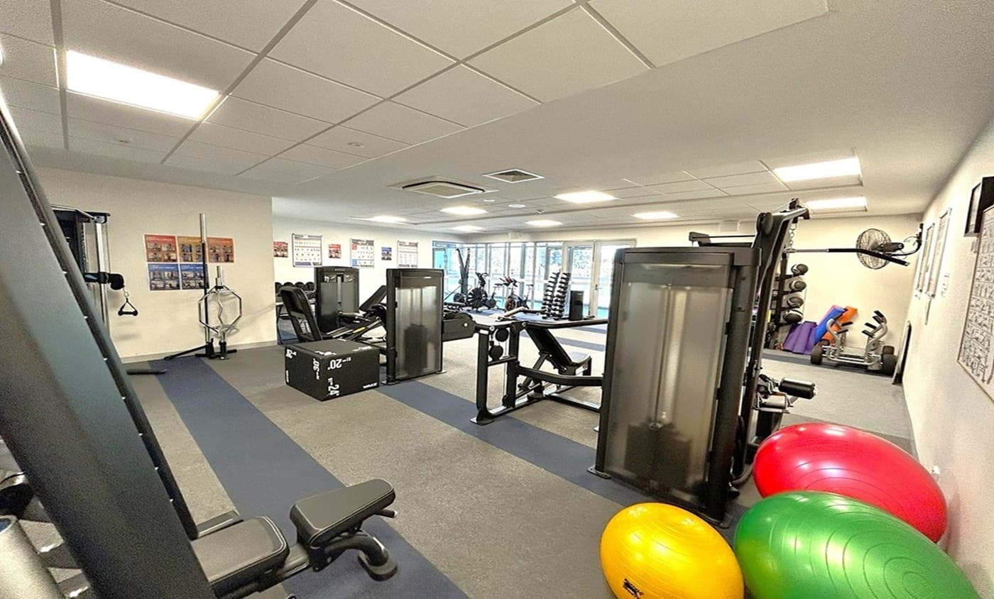 A gym with various exercise equipment. On the left, there are weight machines and a bench. In the centre, a black plyometric box is visible. On the right, there are colourful exercise balls in red, yellow, and green. The background features a wall with posters and a rack of dumbbells. The floor is covered with grey and black mats, and the ceiling has recessed lighting panels.