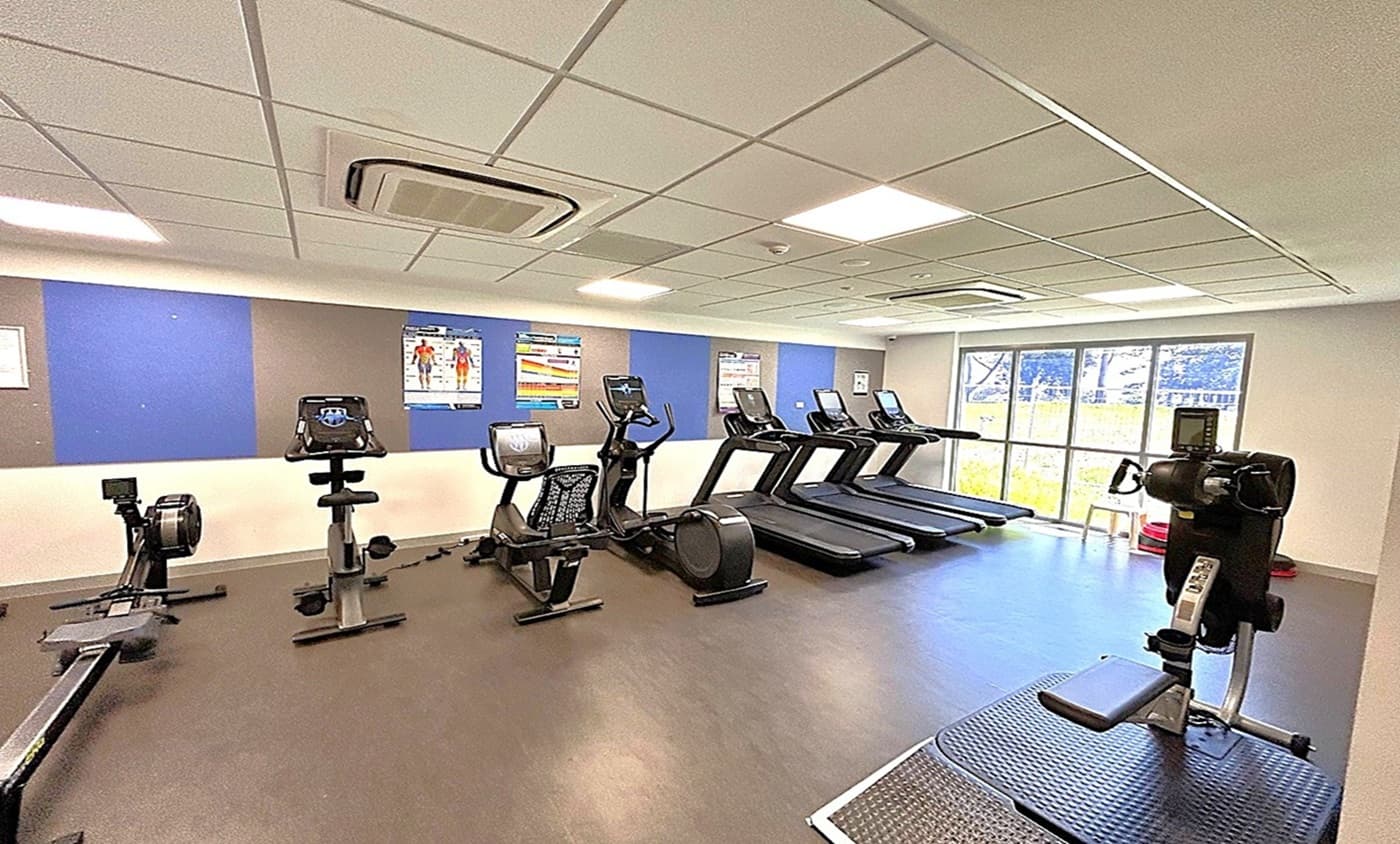 A gym with a variety of exercise equipment. On the left, there are rowing machines and stationary bikes. In the middle, several treadmills are lined up facing large windows that let in natural light. On the right, there's a piece of equipment for upper body workouts. The floor is dark and smooth, and the ceiling is fitted with white tiles and recessed lighting. The walls have blue and grey panels with fitness posters.