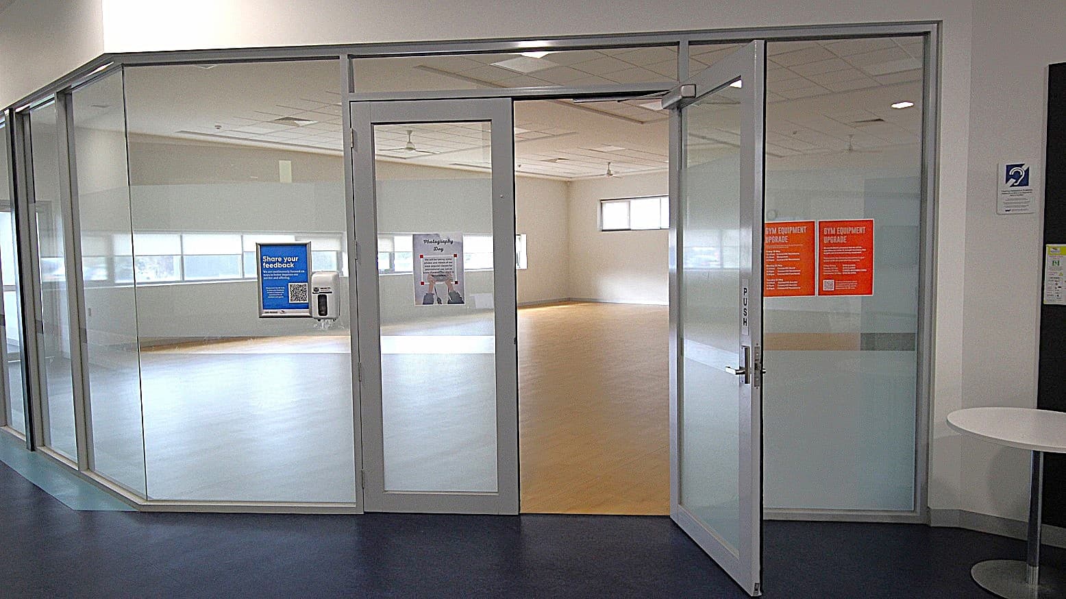 Glass-walled room with a partially open door. The door has a "PUSH" sign and displays a blue feedback poster with a QR code. Inside, the room is empty with light-coloured flooring and several windows allowing natural light. On the right, two orange notices are visible on the glass. A small round table is positioned outside the room on the right. The ceiling has recessed lighting, and the floor outside is dark.