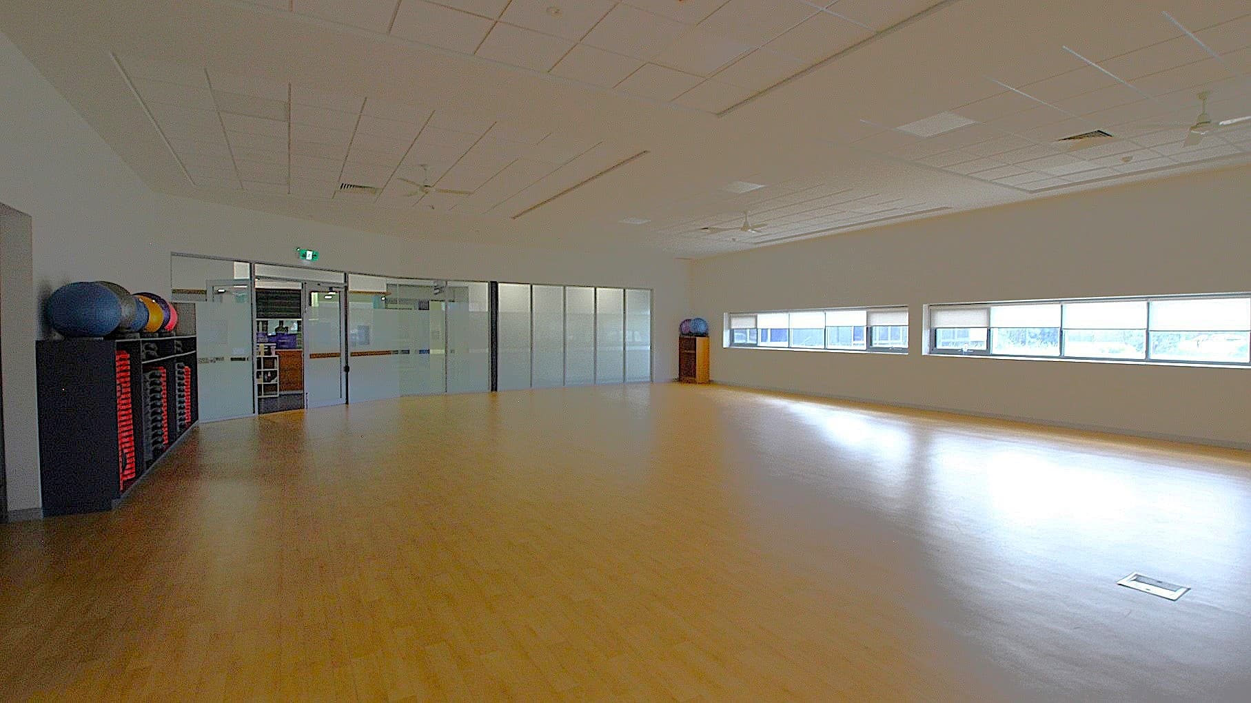 A spacious room with a light wooden floor and a white ceiling featuring recessed lighting and ceiling fans. On the left, there is a rack with exercise balls and red mats. The back wall has large glass doors and windows, allowing natural light to enter. On the right, there are more windows with a view outside. The room appears to be a fitness or dance studio.