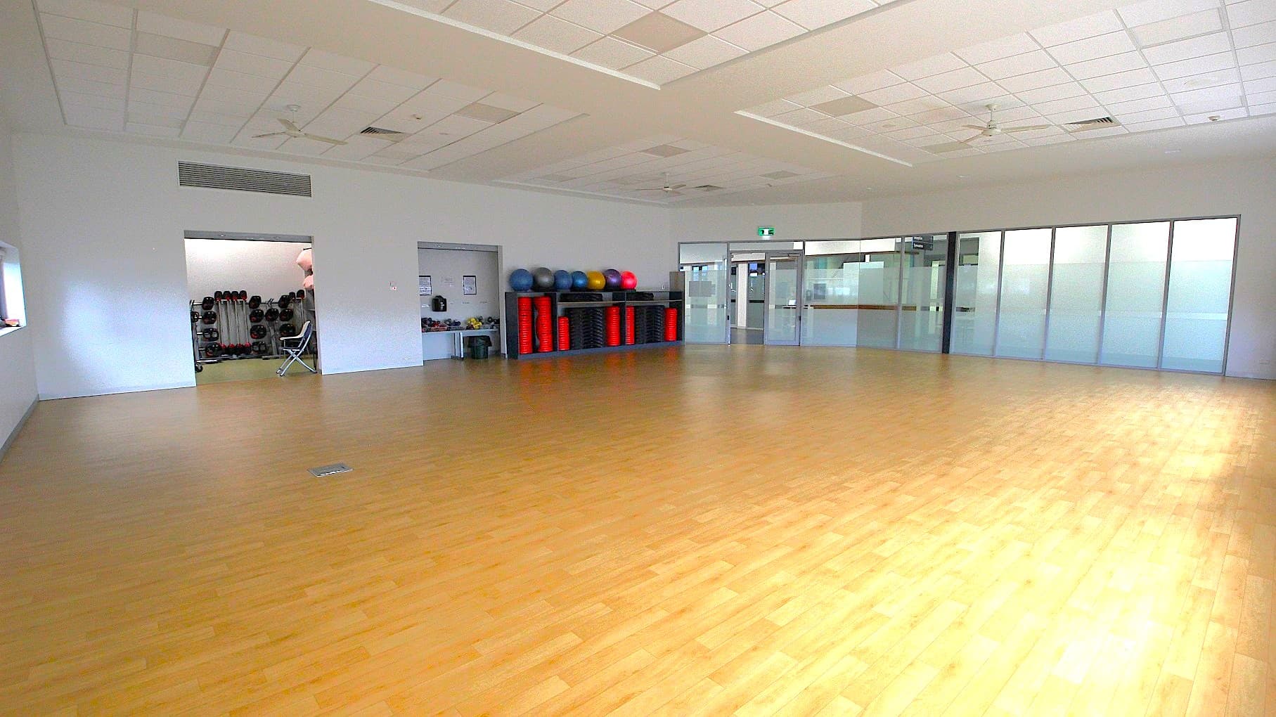 A spacious room with a light wooden floor and a white ceiling with recessed lighting. On the left, there's an open doorway leading to a storage area with exercise equipment like weights and balls. In the centre, a shelf holds various exercise balls and red mats. On the right, there are large glass doors and windows, providing a view into another area. The room is well-lit and appears to be a fitness or multipurpose space.