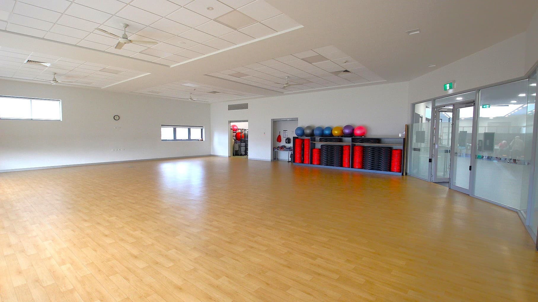 A spacious room with a light wooden floor and a white ceiling featuring ceiling fans. On the right, there are glass doors and a wall with exercise equipment, including colourful exercise balls and stacked red and black steps. The left side has two windows and a clock on the wall. The room is well-lit with natural light coming through the windows.