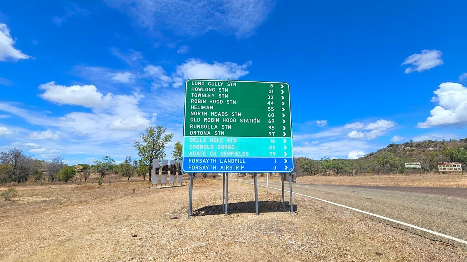 A large road sign stands on a dry, grassy area beside a paved road. The sign lists various destinations with distances in kilometres, including Long Gully Station and Forsayth Airstrip. The background features sparse trees and low hills. The sky is clear with a few clouds. Additional smaller signs are visible further down the road on the right.