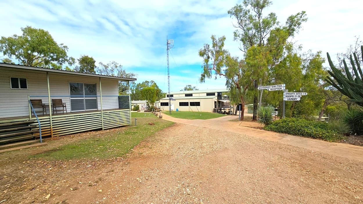 A gravel path leads through a rural area. On the left, a small building with a porch and two chairs is visible. In the centre, a larger building with a metal tower behind it is surrounded by trees. On the right, there are directional signs pointing to various facilities, and a large plant with spiky leaves. The ground is a mix of grass and dirt, and the area is dotted with trees.