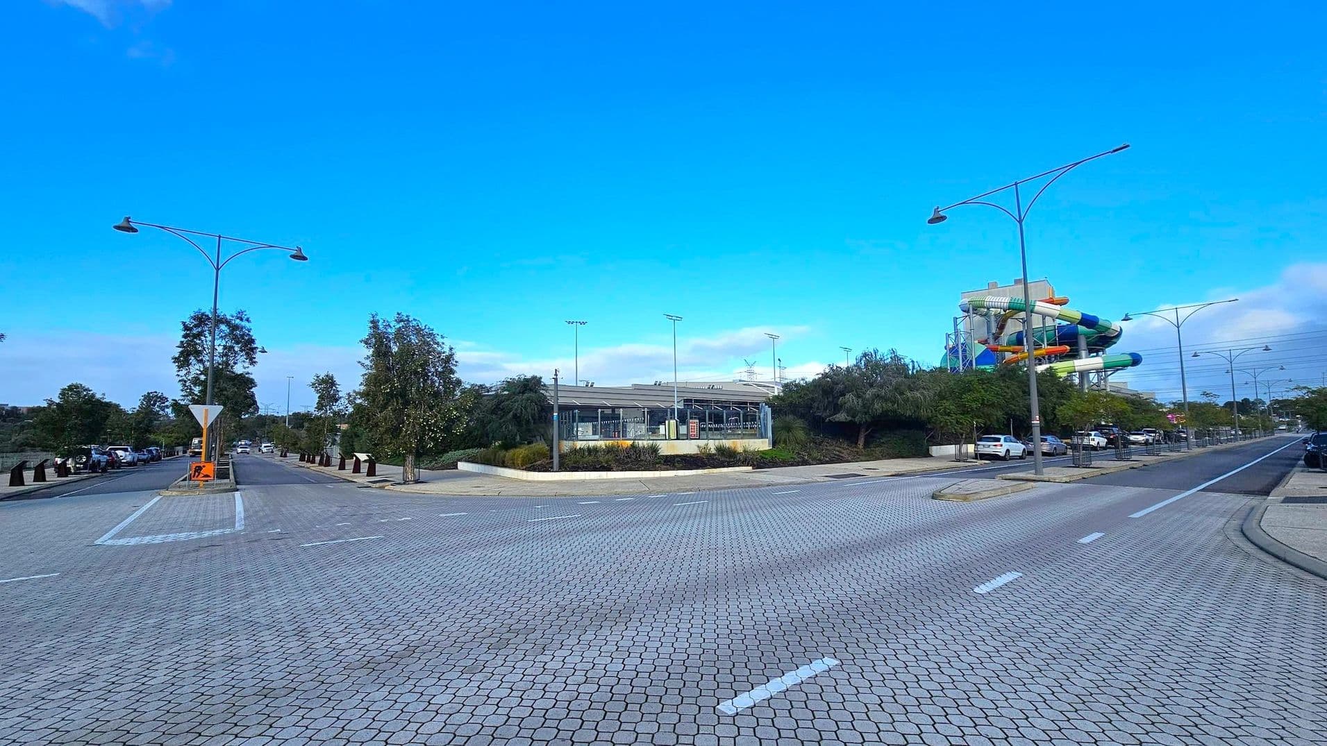 A wide, paved road with a textured surface runs through the foreground. On the left, there are trees lining the street and parked cars. In the centre, a building with a modern design is visible, surrounded by greenery. On the right, a colourful water slide structure is prominent, with more parked cars and trees. Tall streetlights line both sides of the road.