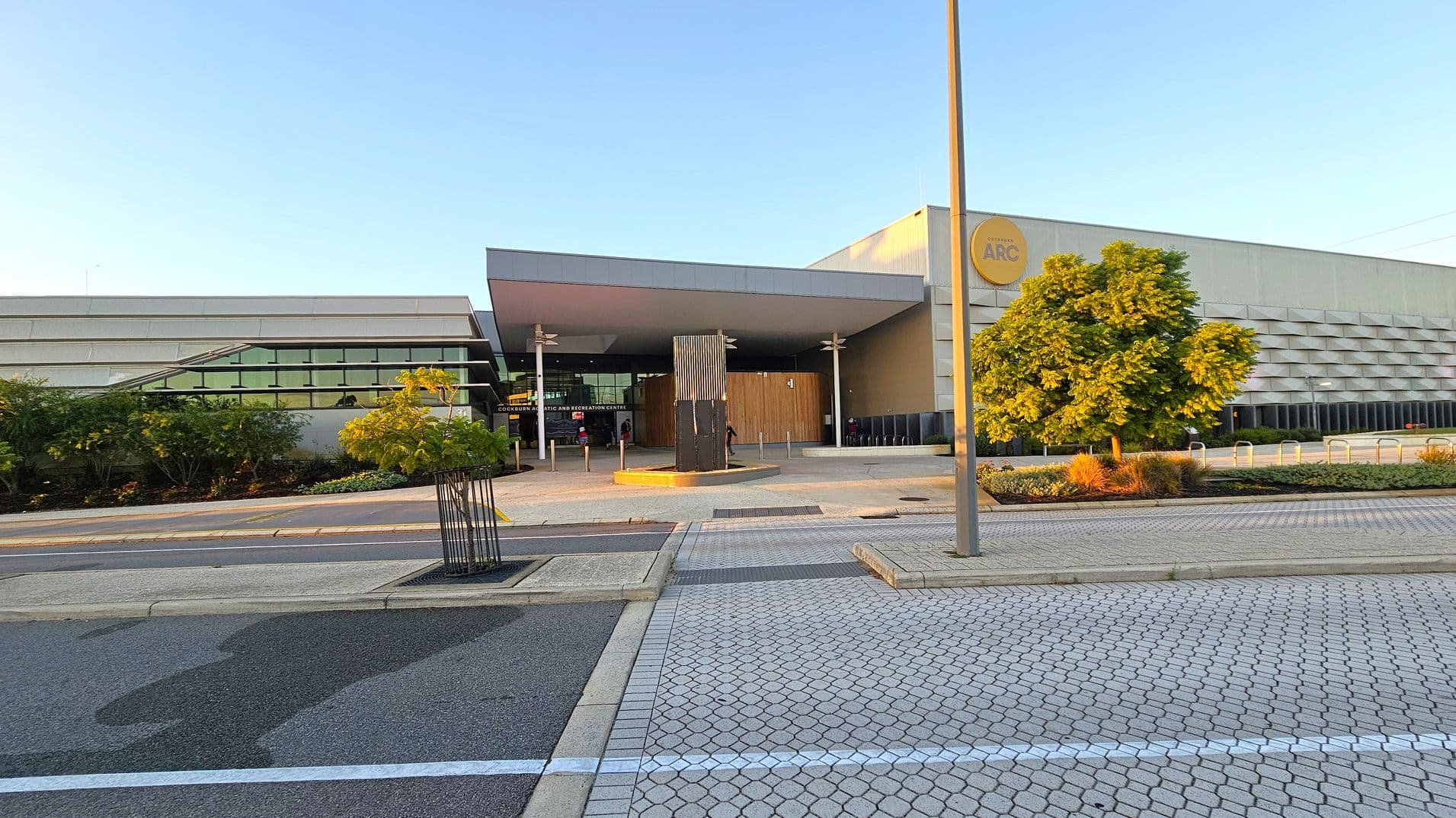 A modern building with a large entrance is in the centre, featuring a wooden panelled section and a sign reading "ARC". On the left, there are horizontal windows and landscaped greenery. On the right, a tree and more greenery are visible. The foreground has a paved area with a mix of hexagonal and rectangular patterns, and a street lamp is present. The building has a flat roof and a sleek, contemporary design.