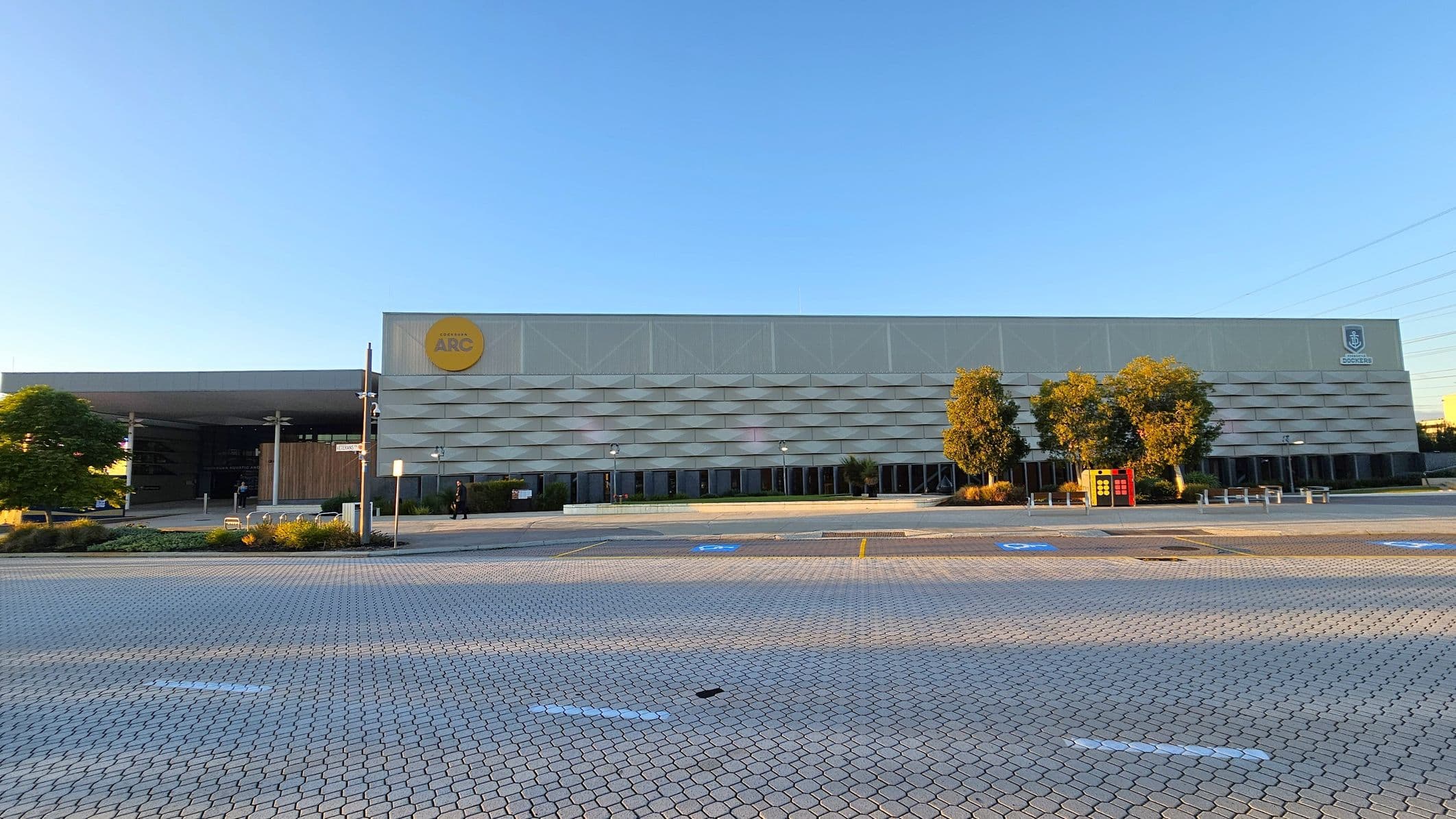A large, modern building with a textured facade and a yellow circular sign on the left side. The entrance is partially visible with a wooden panelled section. Several trees and shrubs are in front of the building, along with a red vending machine. The foreground features a paved area with parking spaces marked, including accessible parking spots. The sky is clear, and there are no visible people.
