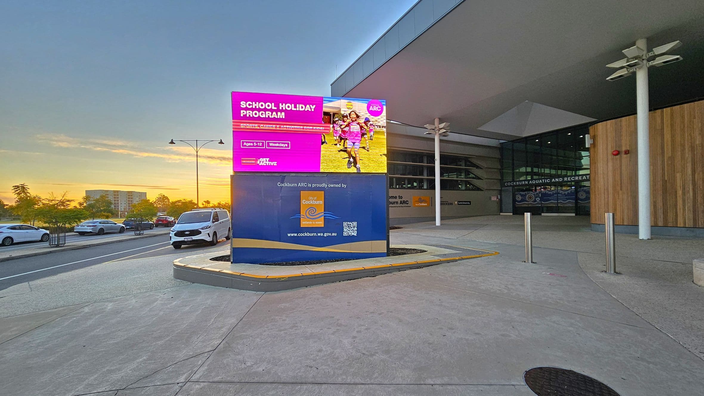 On the left, several cars are parked along a road with trees and a building in the background. In the centre, a large digital sign displays a "School Holiday Program" advertisement with children running. On the right, the entrance to the Cockburn Aquatic and Recreation Centre features glass doors and wooden panelling. The foreground has a concrete pavement with metal bollards, and the ceiling above the entrance is angular and modern.